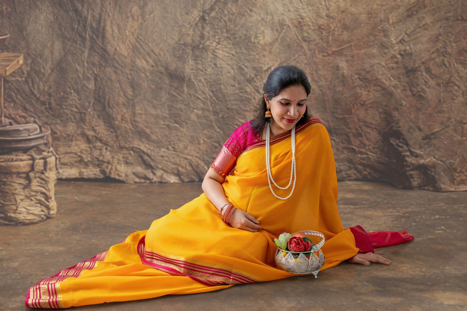 Woman in yellow and pink saree with white pearl necklace sitting gracefully on floor holding decorative pot.