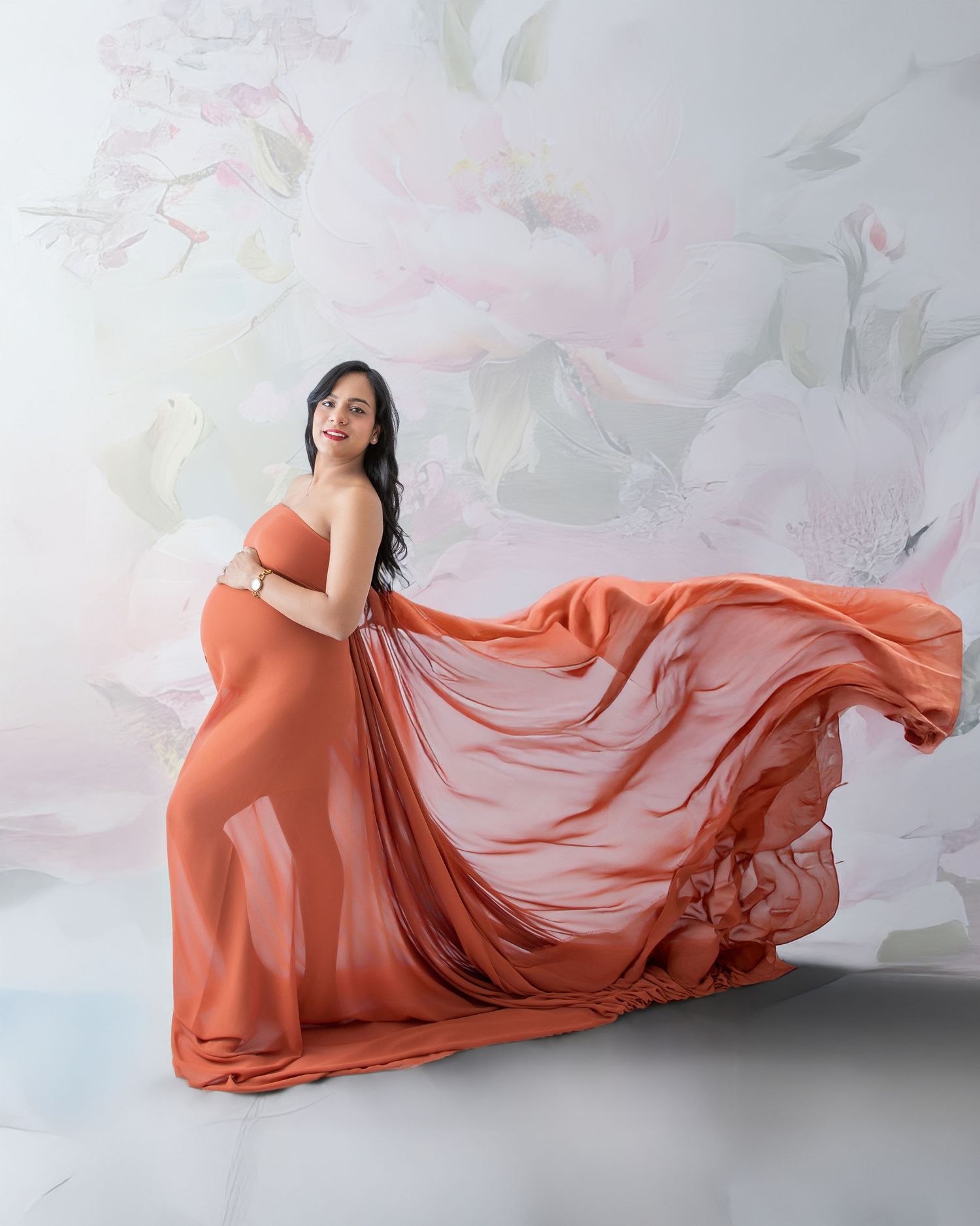 Pregnant woman in flowing orange maternity gown poses gracefully against a soft floral backdrop in a studio.