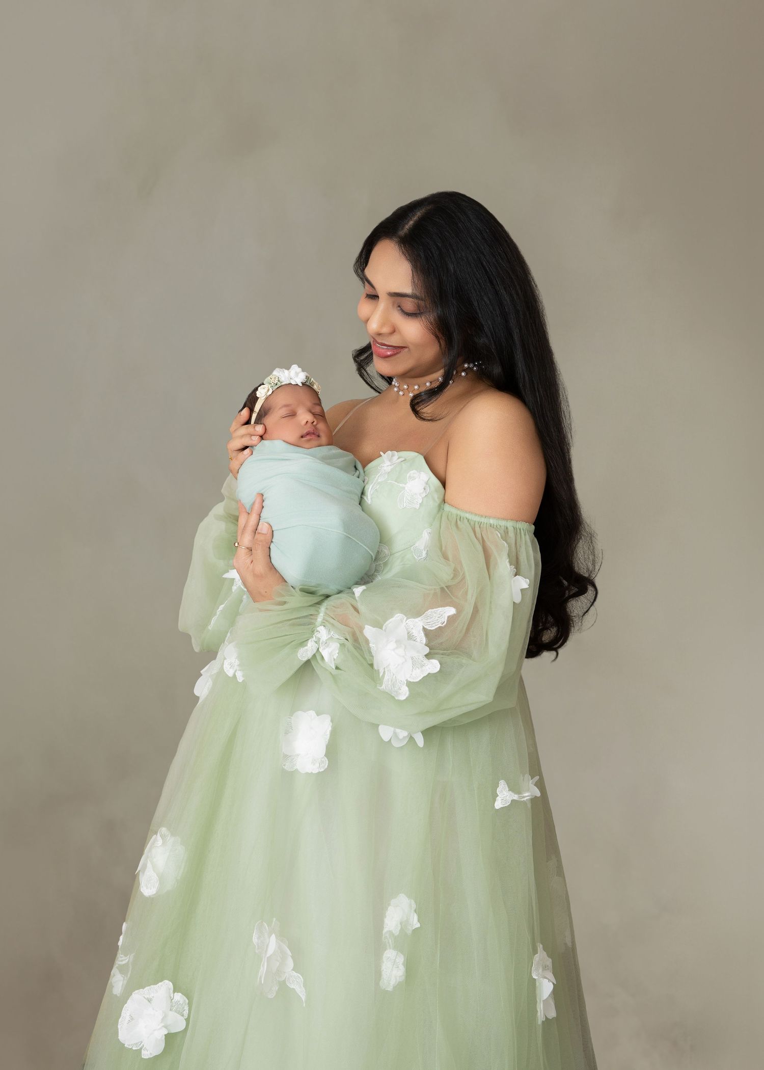 A woman in a sage green floral gown holds her swaddled newborn baby during a professional maternity photo session.