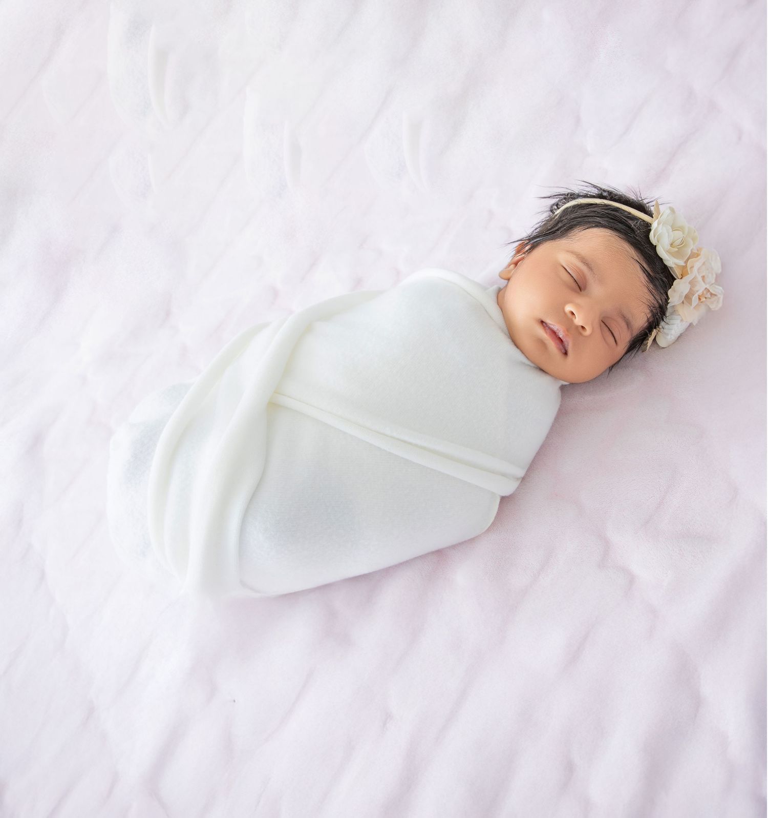 Sleeping newborn baby swaddled in white blanket on soft white surface with delicate headband.