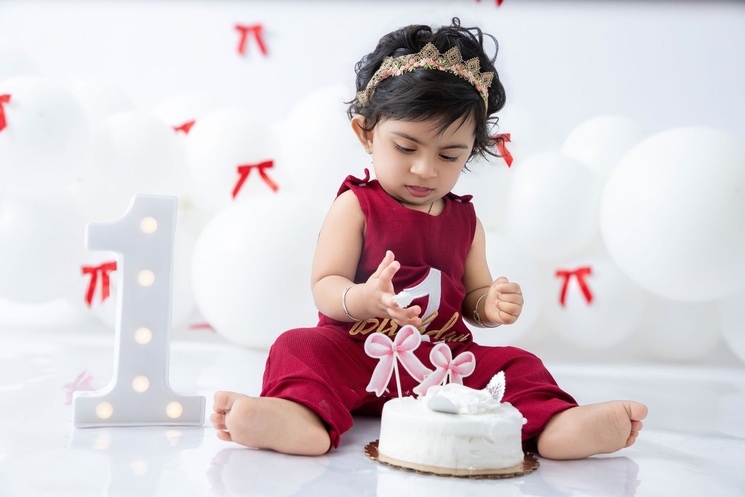 Baby reaching for white cake during first birthday celebration with red bow decorations.