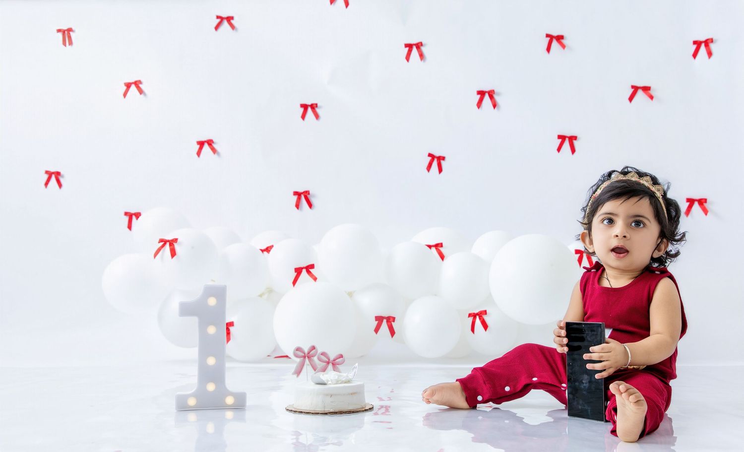 Baby in red outfit explores white decorated space with number one decor.