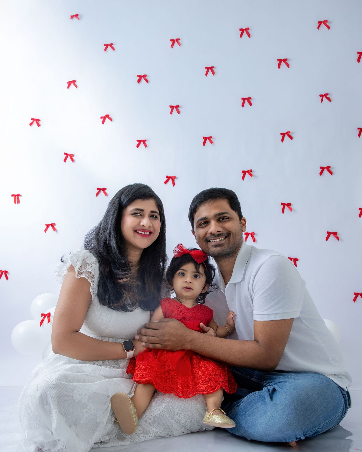 A family poses together against a white backdrop with red bows, wearing coordinated white and red outfits.