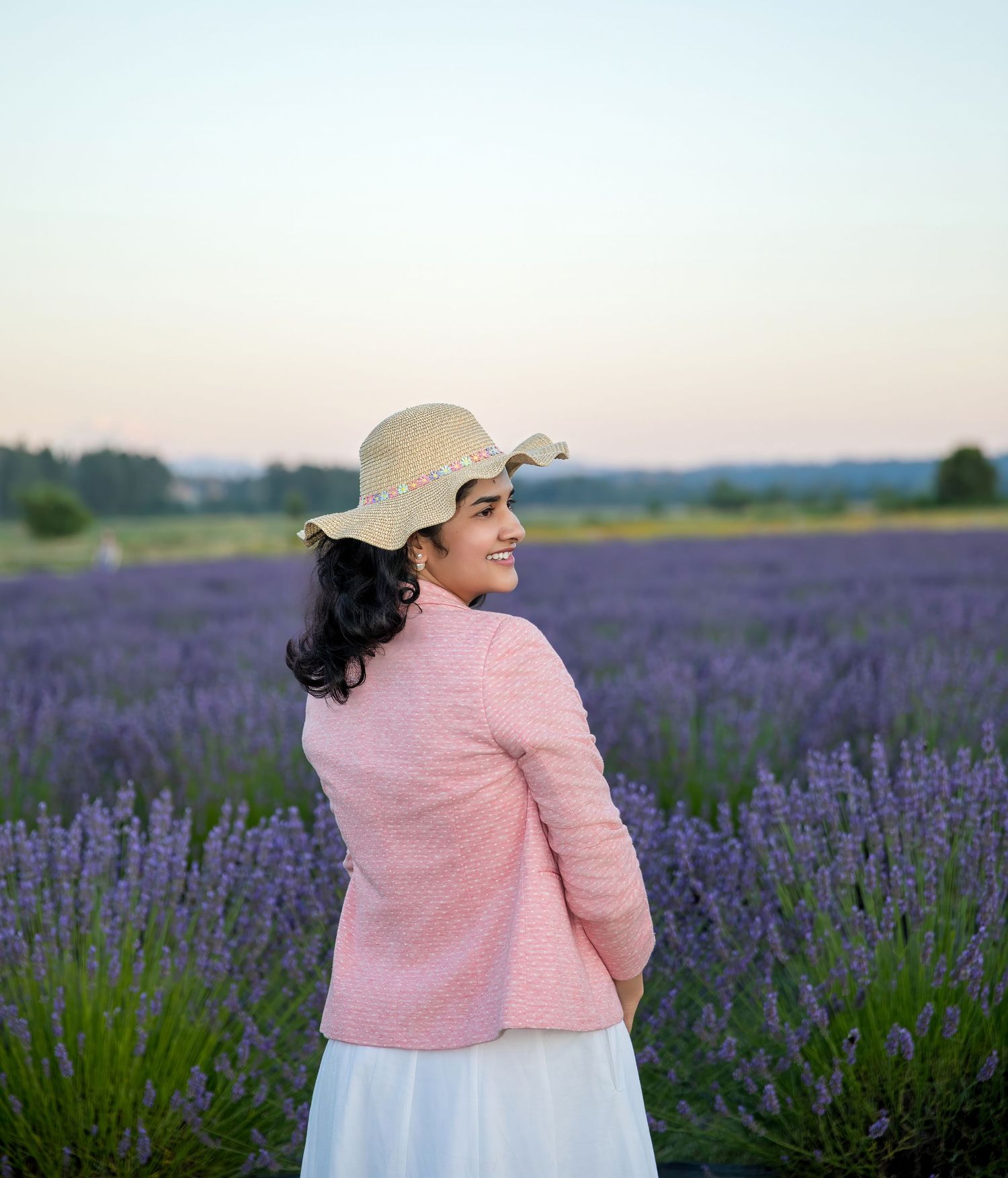 Person wearing pink jacket and sun hat poses in lavender field at dusk.