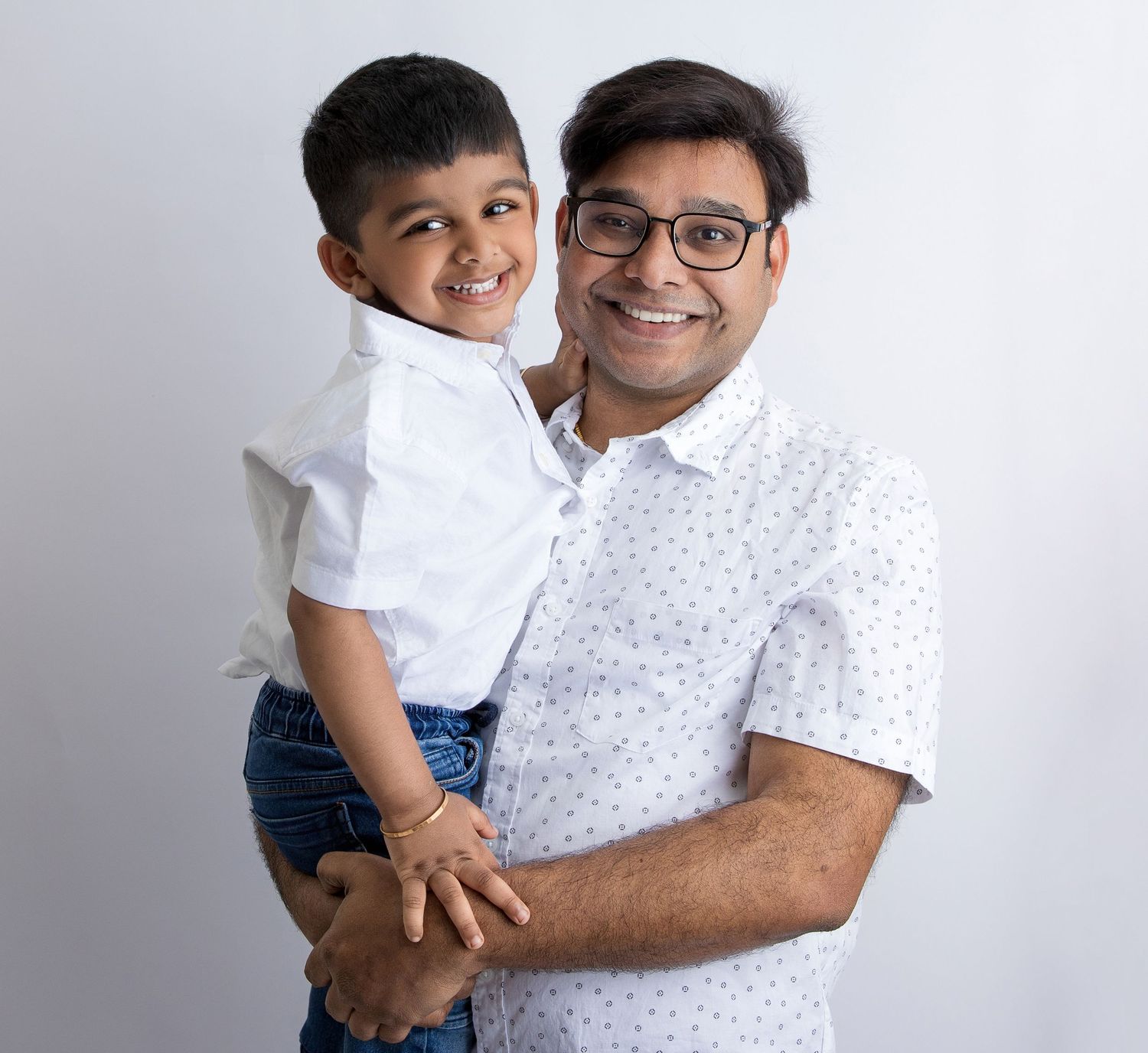 Father and young child pose together in matching white shirts against a light background.