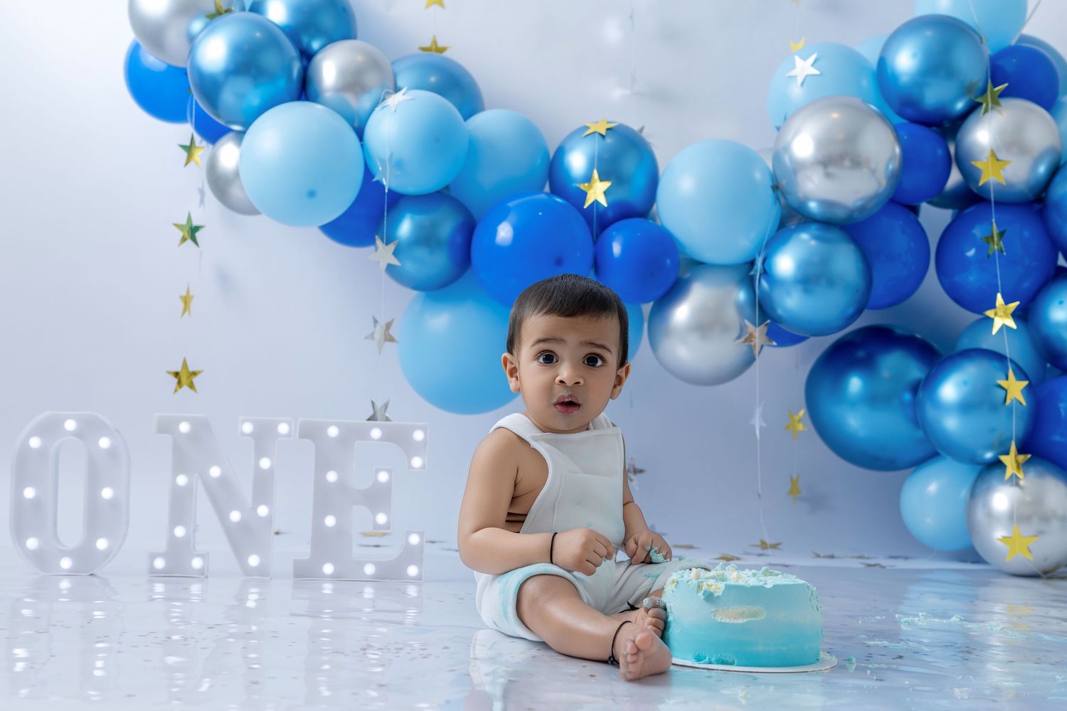 A baby in white sits on a floor with blue and silver balloon decorations for a first birthday celebration.