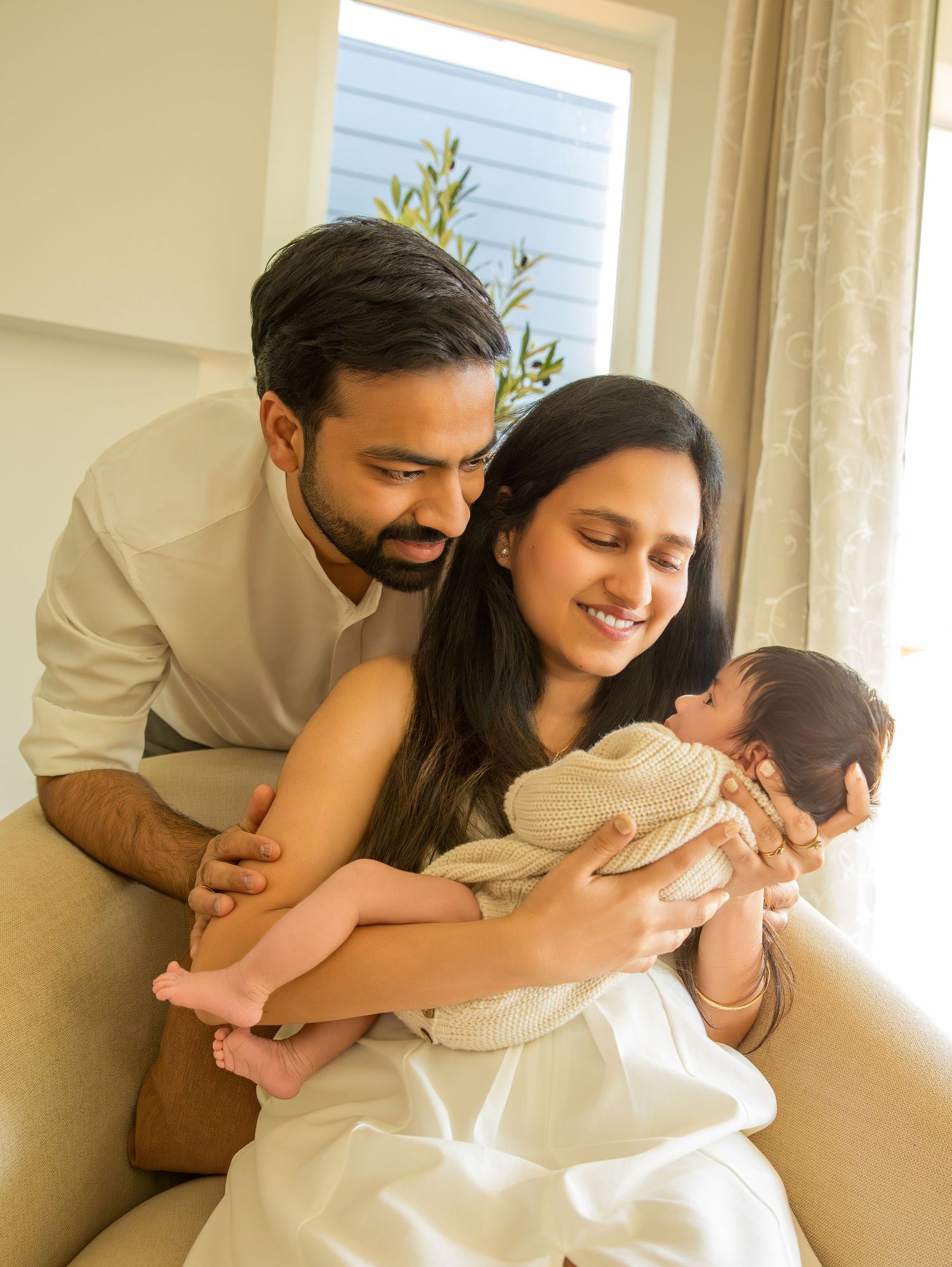 Loving family photo sequence showing parents embracing their newborn on a couch near a window.