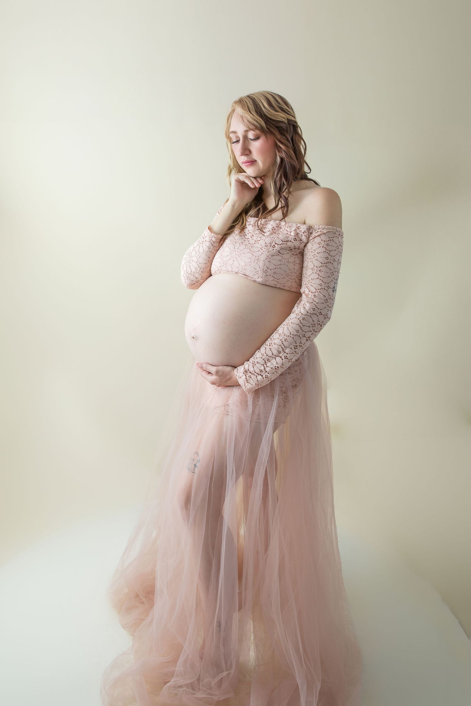 Pregnant woman in blush pink lace and tulle maternity gown cradling her baby bump in a softly lit studio.