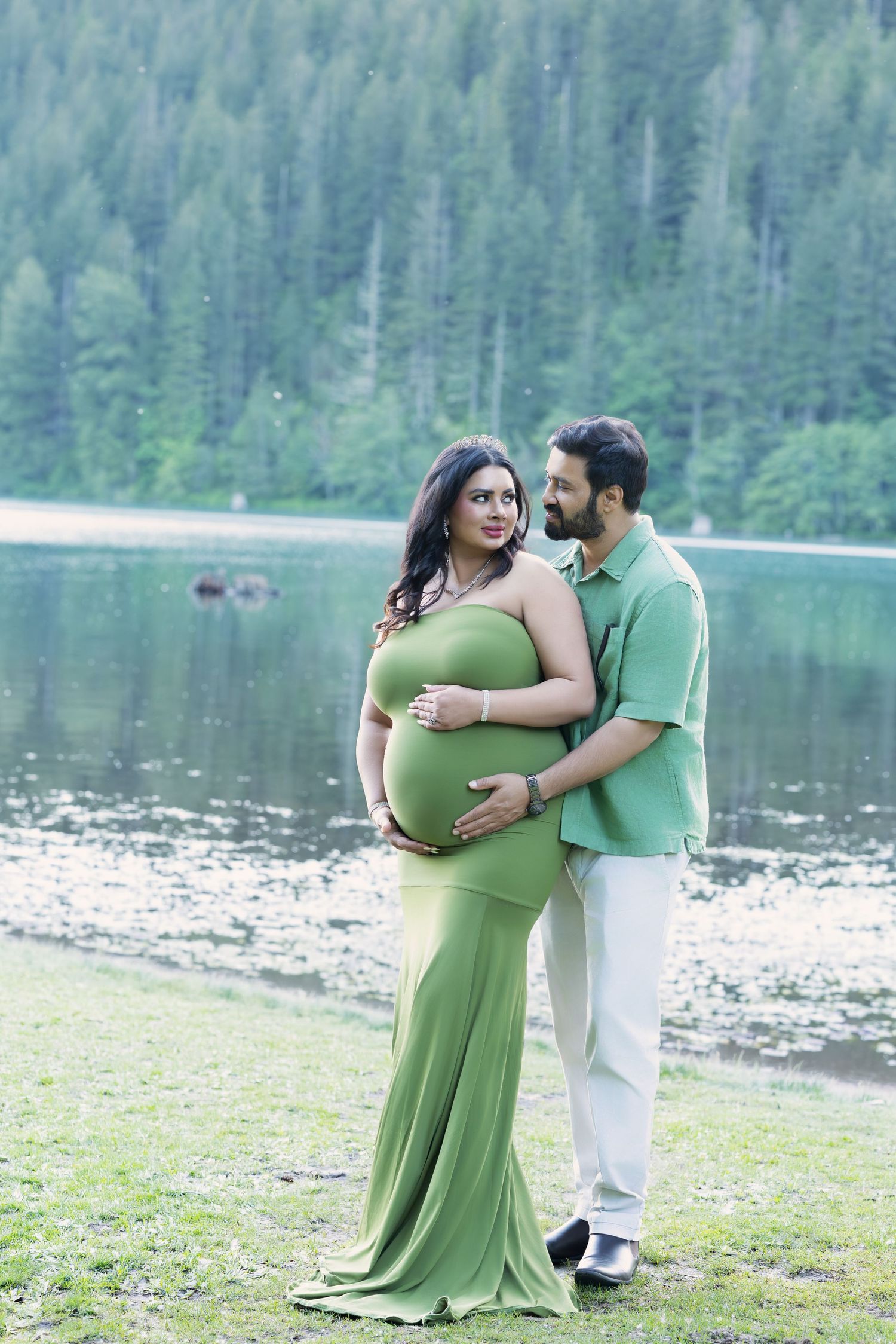 Pregnant woman in green gown embraced by partner near a serene lake with forested hills backdrop.