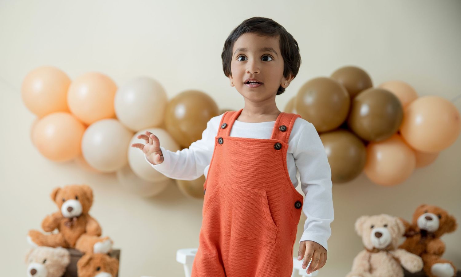 Child in orange overalls standing in front of balloon arch and teddy bear display.