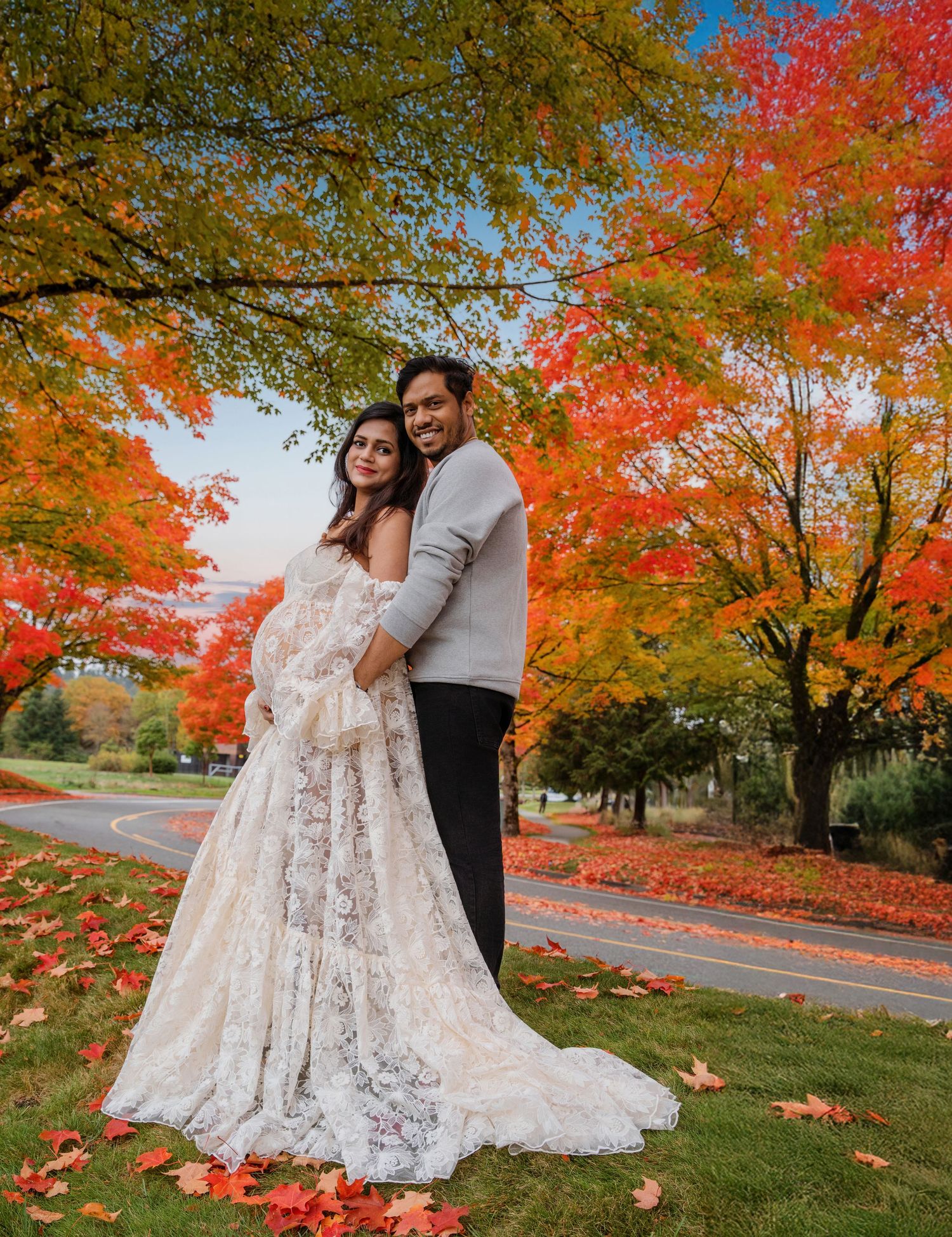 Couple poses in autumn park, woman in lace maternity gown, man in gray sweater, vibrant red and orange fall foliage.