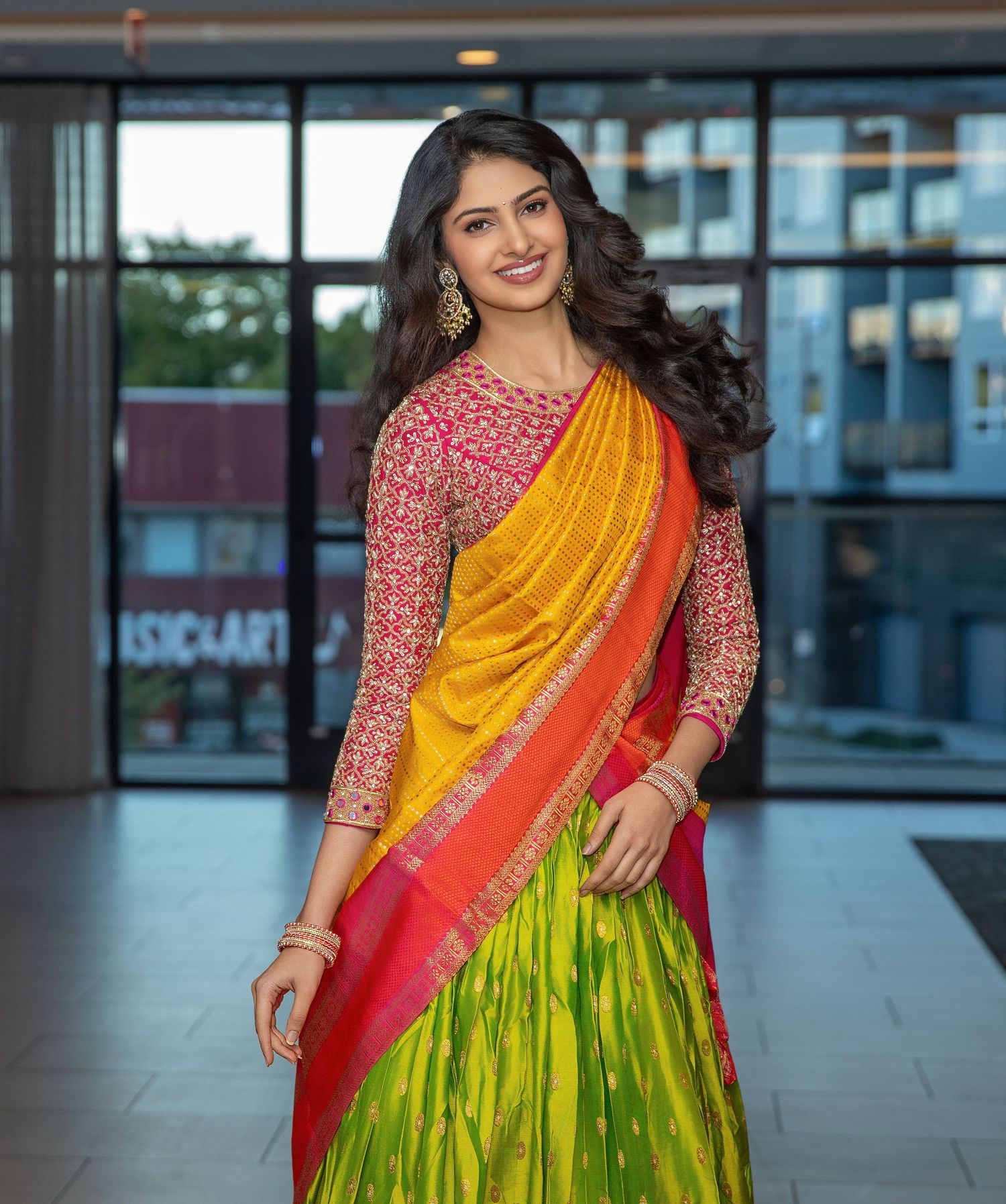 A woman wearing a vibrant yellow and pink silk saree with a green pleated skirt poses elegantly in an indoor setting.