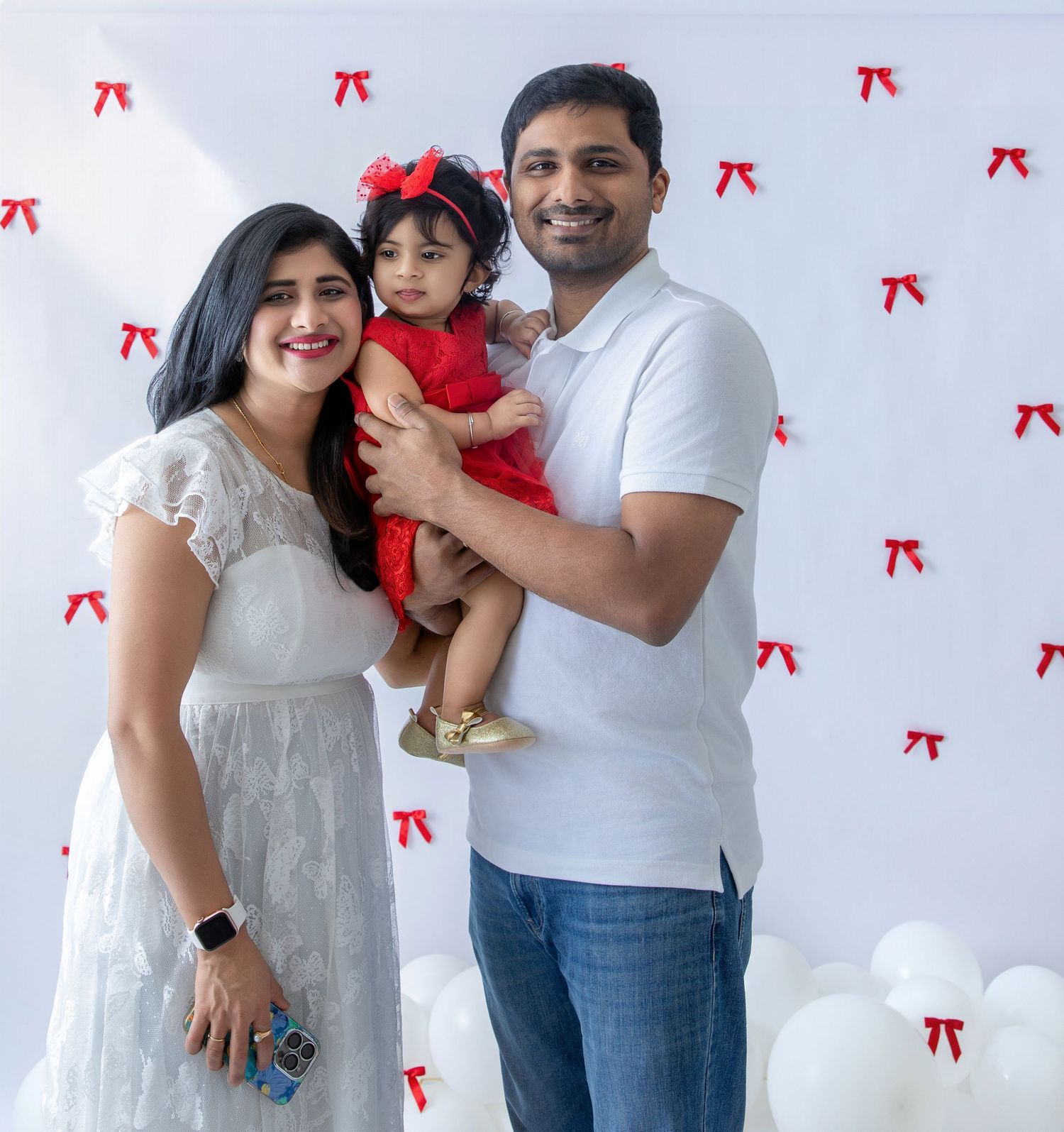A family poses together against a white wall decorated with small red bows in a holiday photoshoot setting.