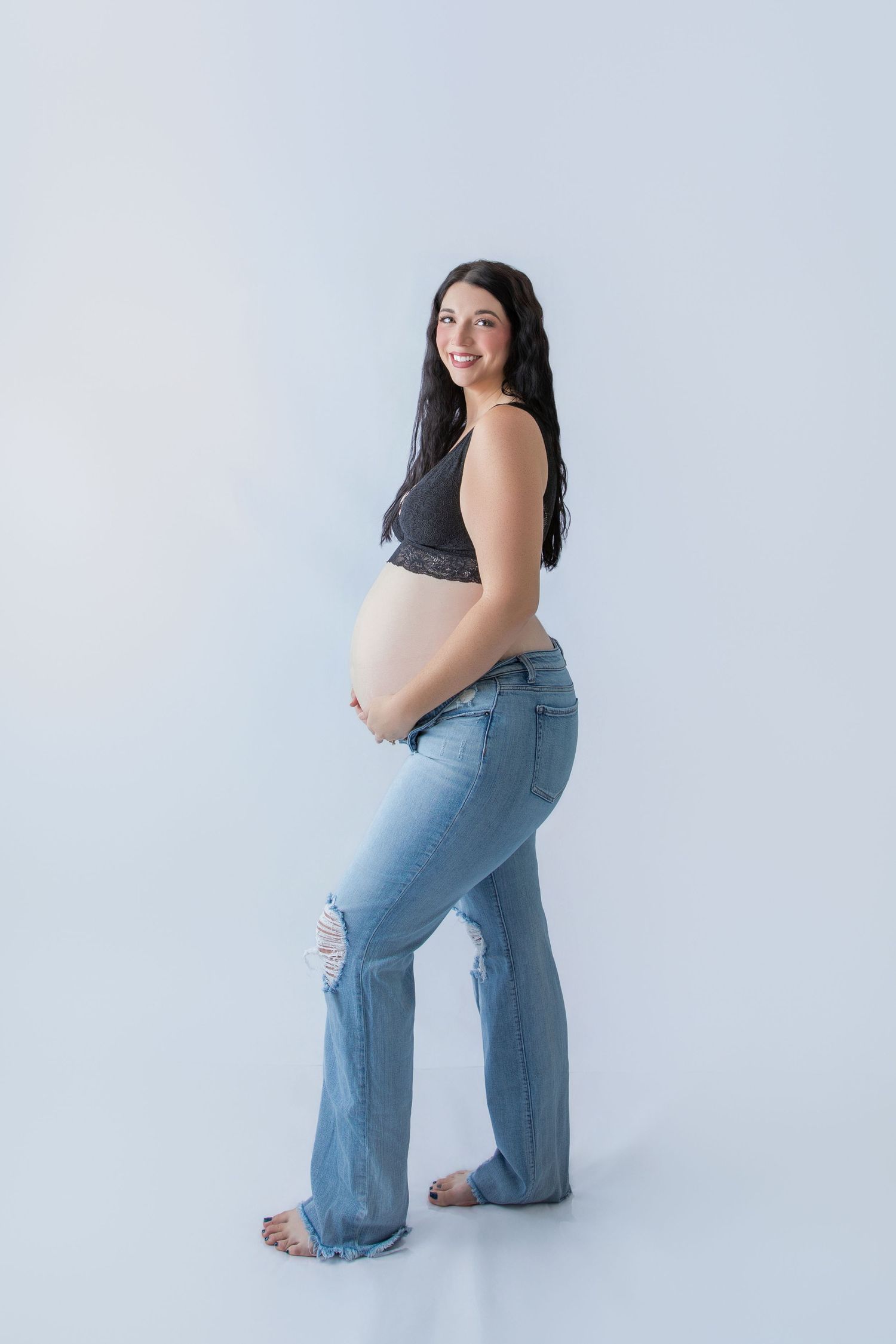 Smiling pregnant woman in ripped jeans and black lace bralette cradling her baby bump against a white background.