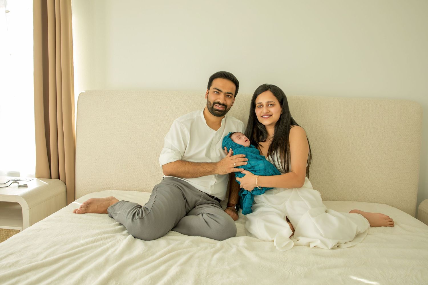 A couple relaxes and laughs together while sitting in bed with white bedding and beige curtains in the background.