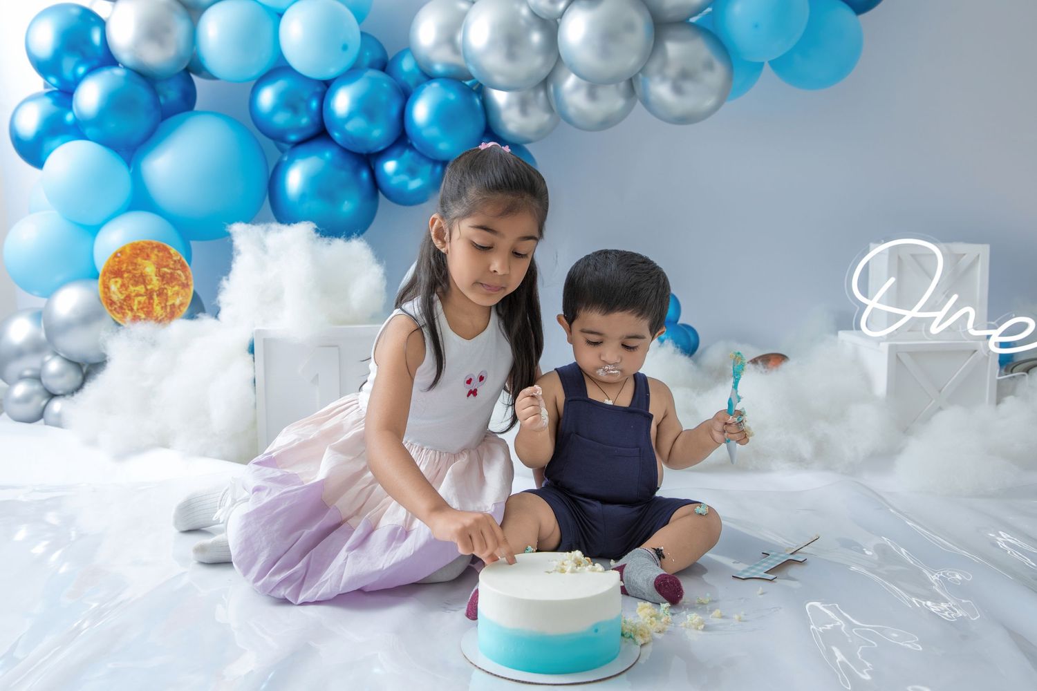 Two children sit on a white floor with a blue and white cake surrounded by balloon decorations.