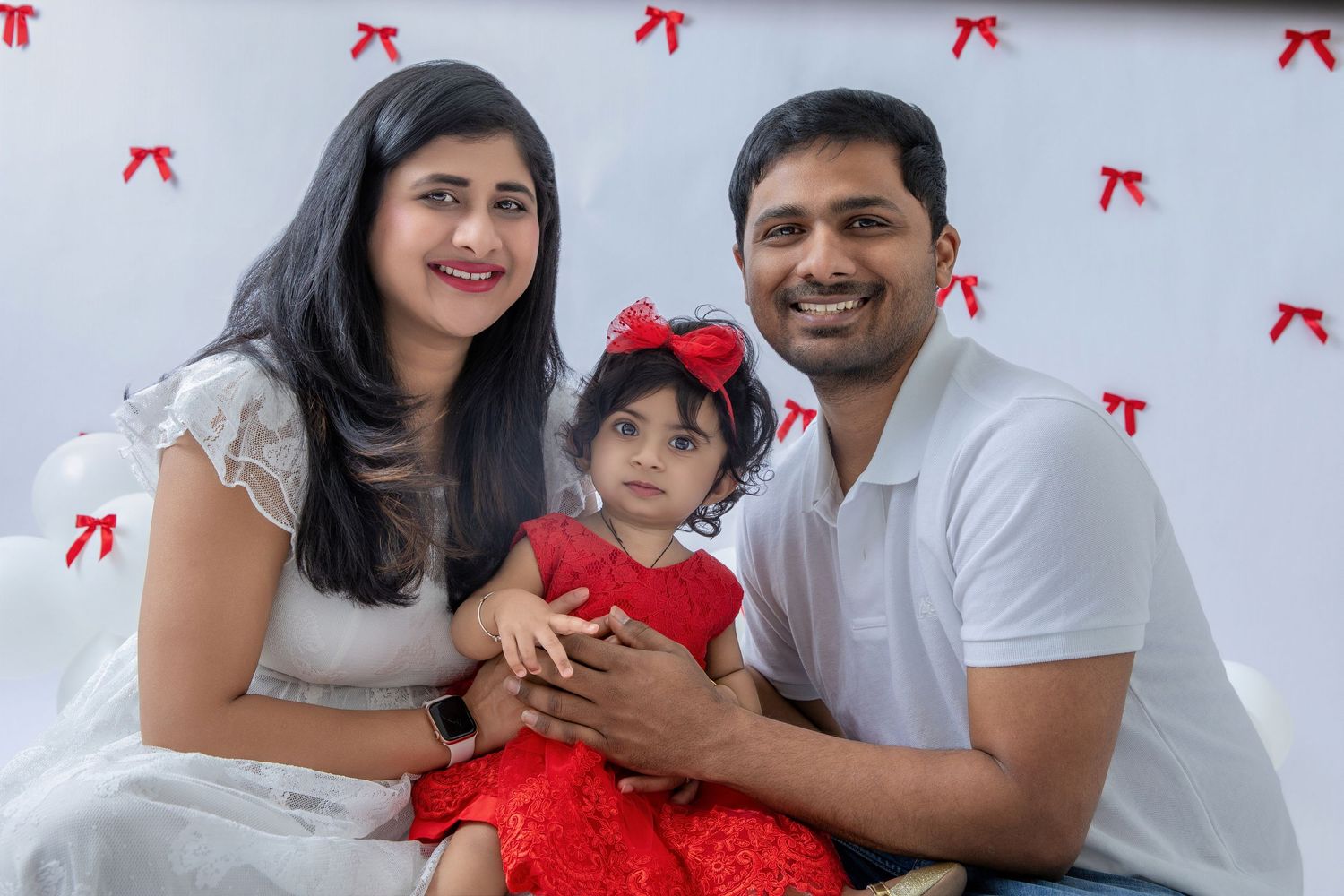 Parents and child in coordinated white and red outfits pose for holiday photos against decorated wall.