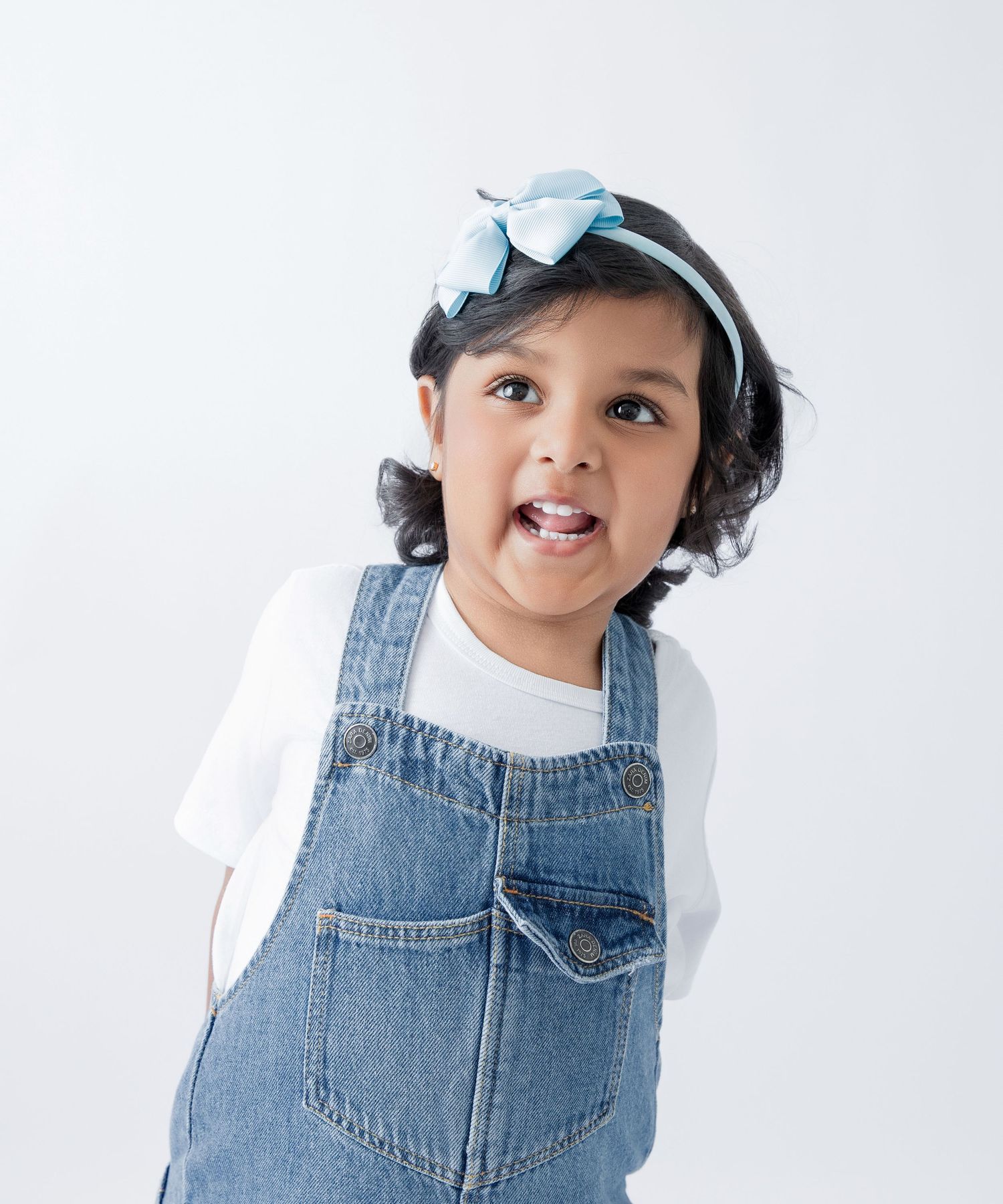 Child wearing denim overalls and white shirt poses playfully against white background.
