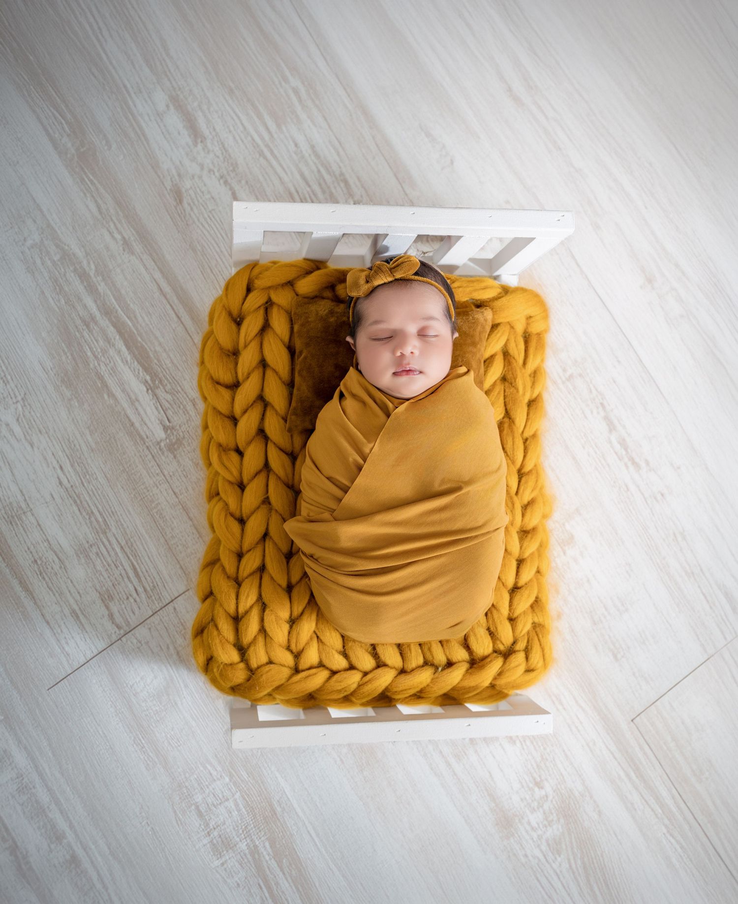 Newborn baby swaddled in mustard yellow wrap sleeping on chunky knit blanket in white mini crib.