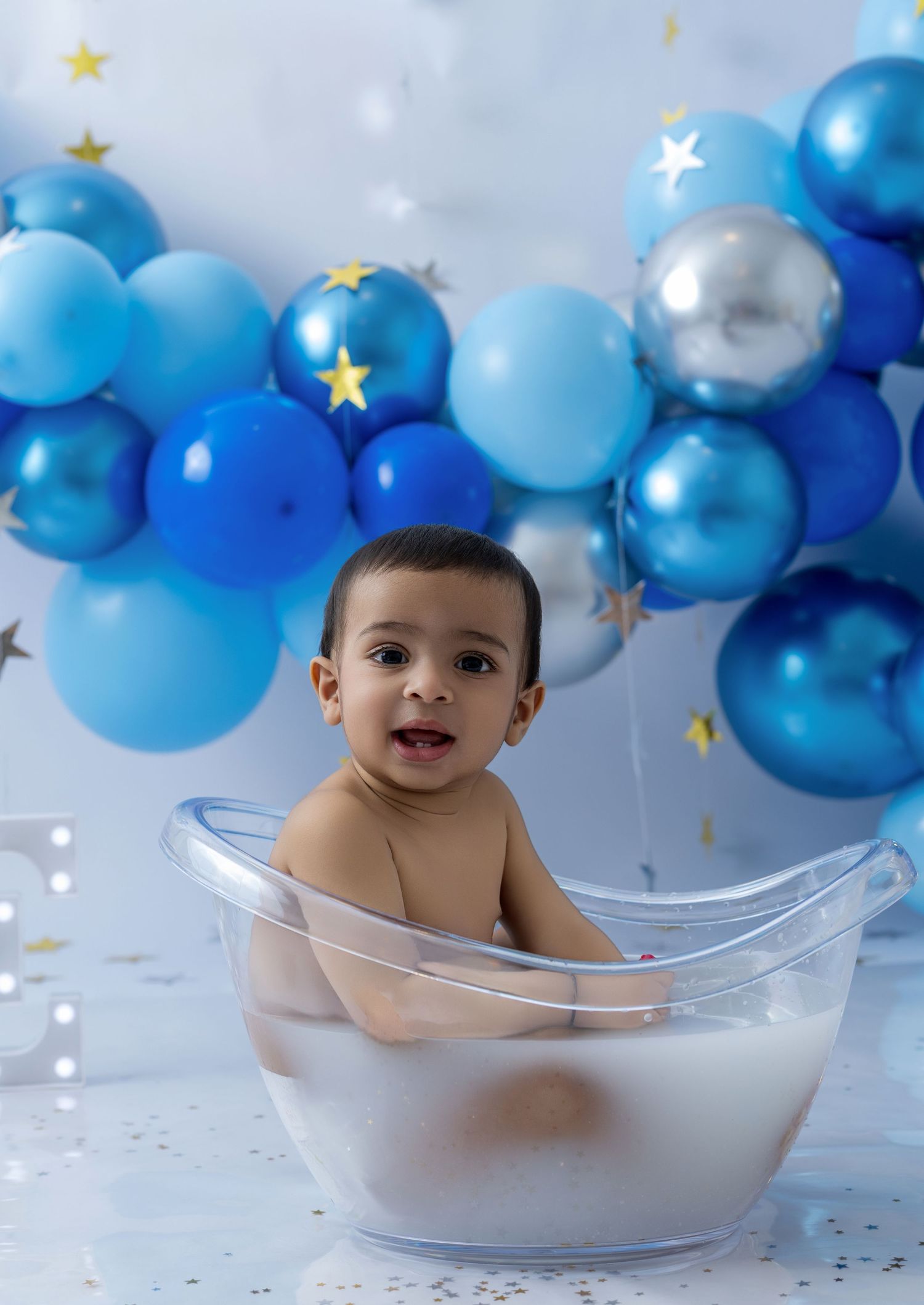 Baby sits in clear bathtub with blue and silver balloons and star decorations for milestone photo shoot.