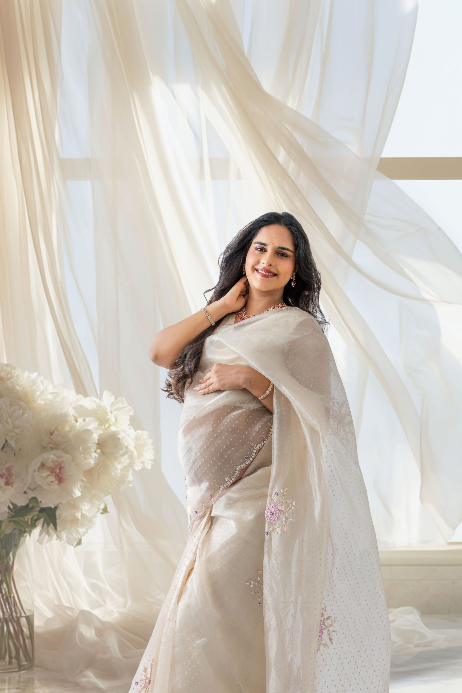 A woman in an elegant cream embroidered saree poses gracefully near sheer white curtains with soft natural light.