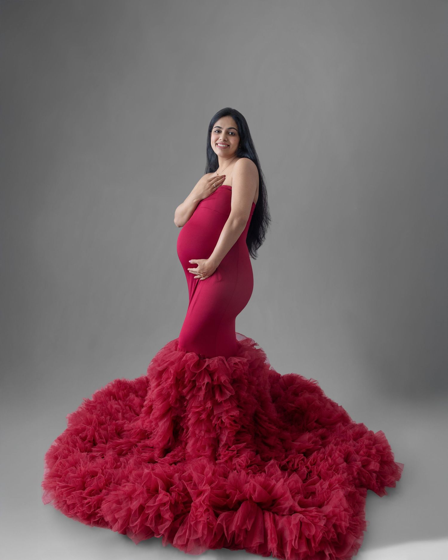 Pregnant woman in elegant red mermaid gown with ruffled tulle train poses gracefully in studio maternity photo.