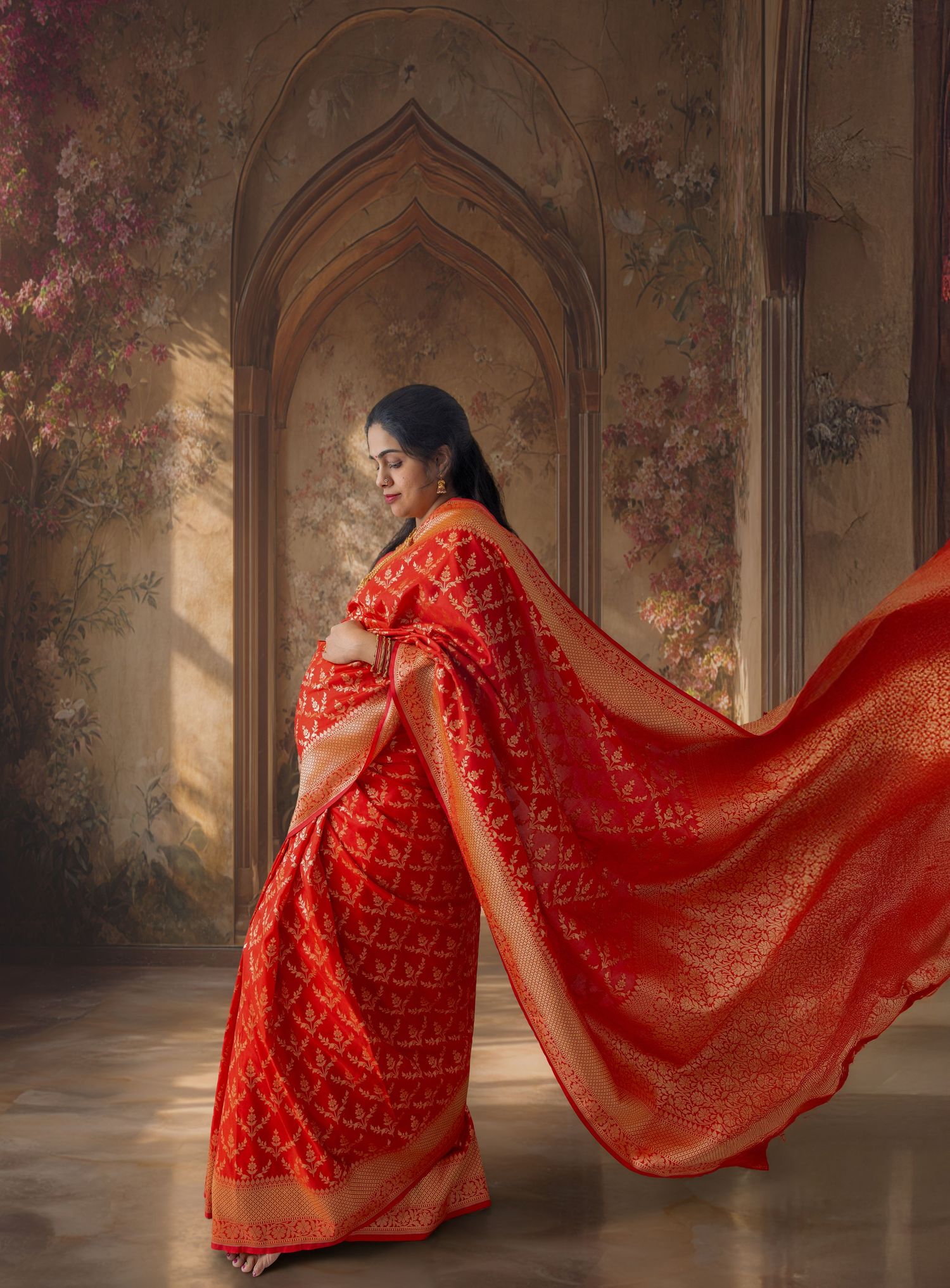 A woman in a flowing red and gold Banarasi saree poses gracefully in an ornate floral archway setting.