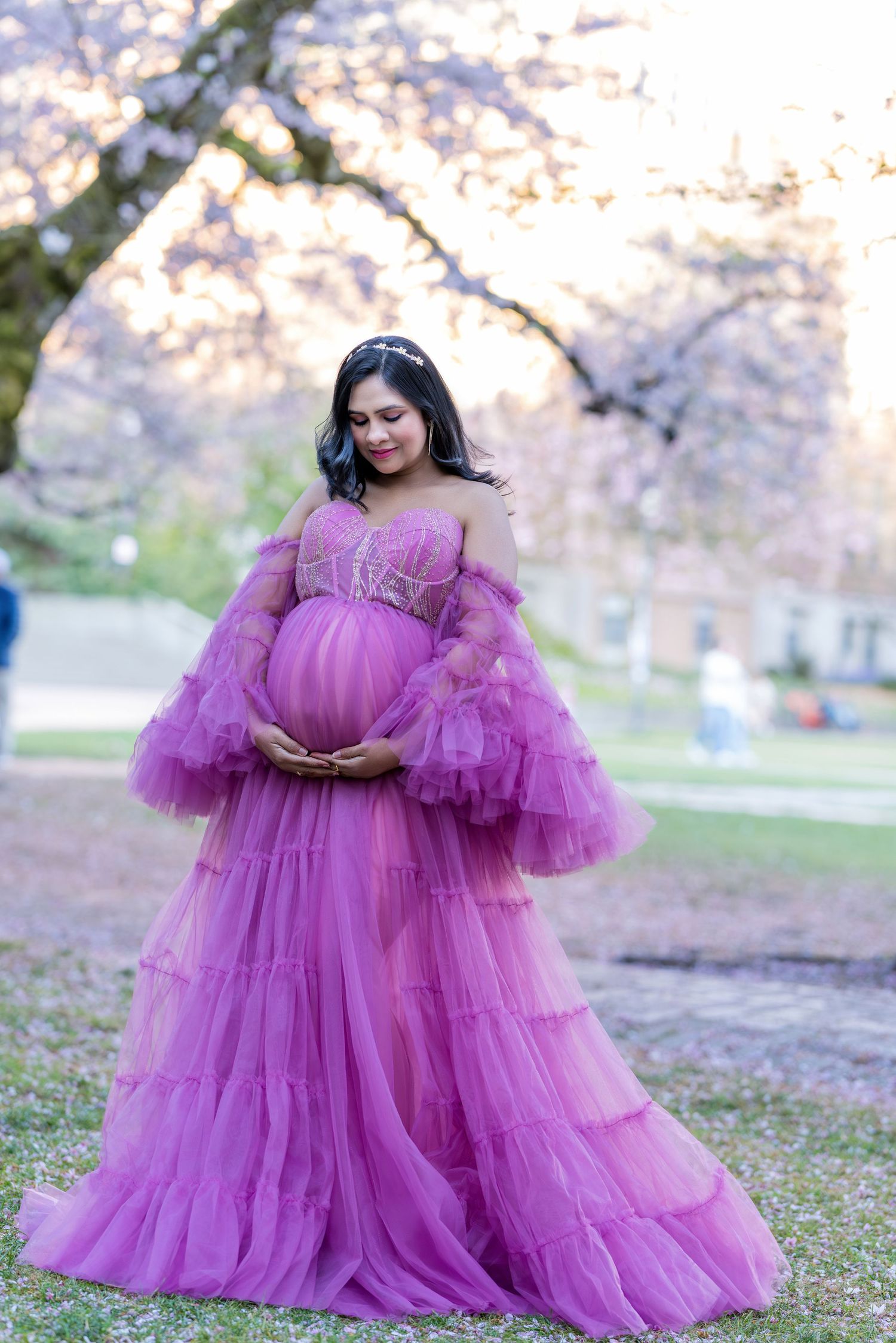 Pregnant woman in stunning purple tulle off-shoulder gown cradling baby bump under cherry blossom trees.