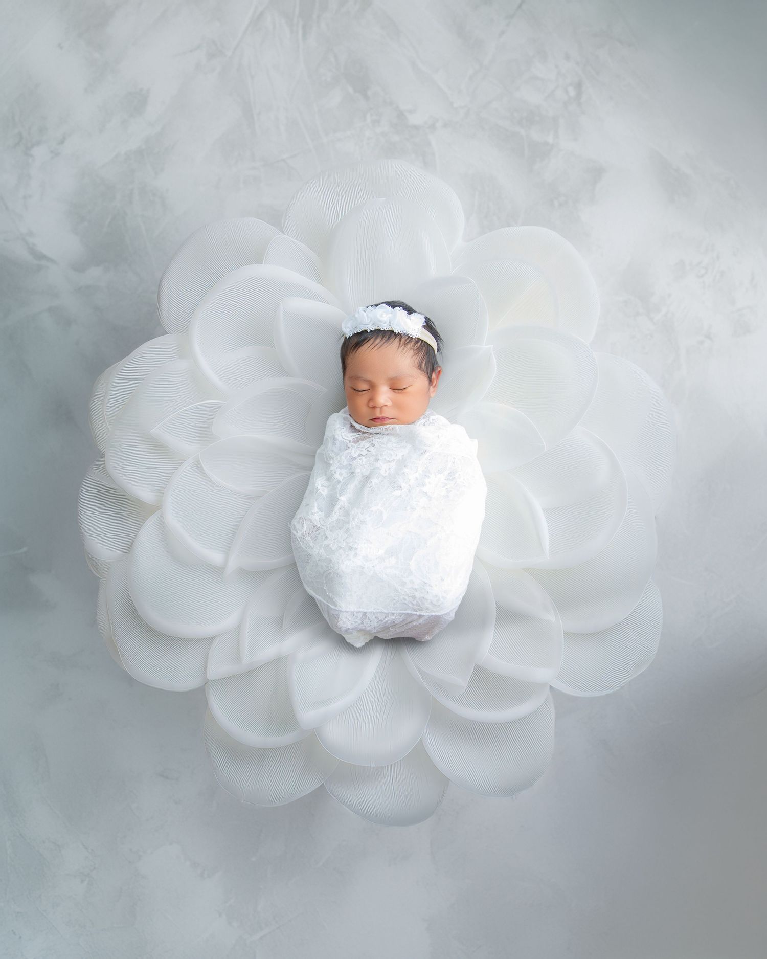 Sleeping newborn baby swaddled in white lace wrap with floral headband, resting on large white flower petals.