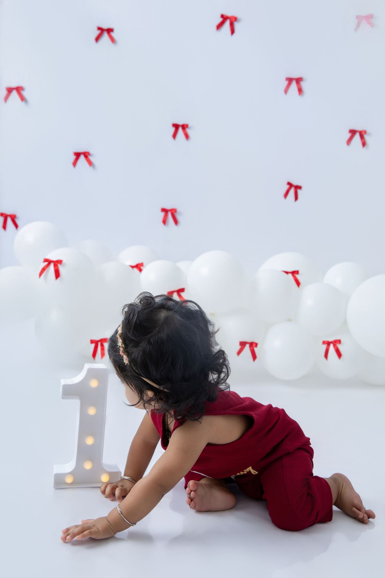 Baby in burgundy outfit crawling near illuminated number one decoration.