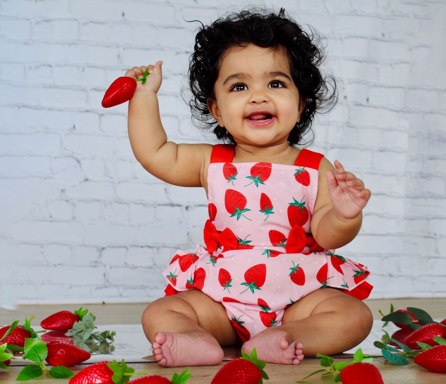 Baby in floral dress surrounded by red roses against white brick background.