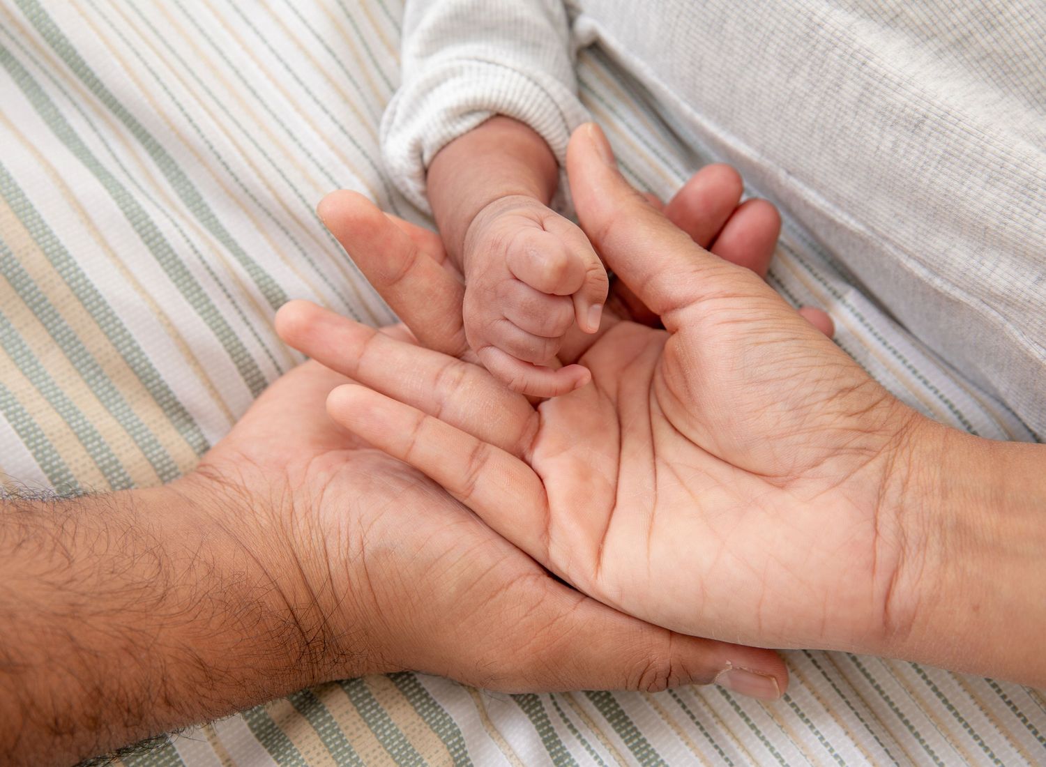 A tiny newborn baby hand resting gently in an adult's open palm on a striped blanket.