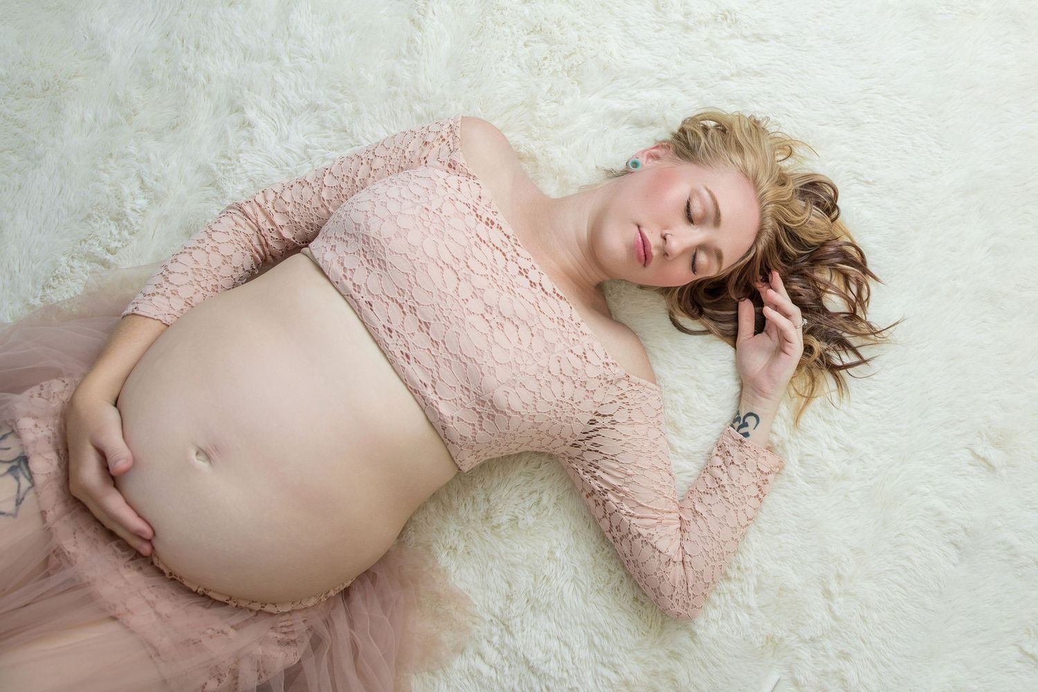 Pregnant woman in pink lace crop top and tulle skirt lying on white fur, cradling her baby bump.