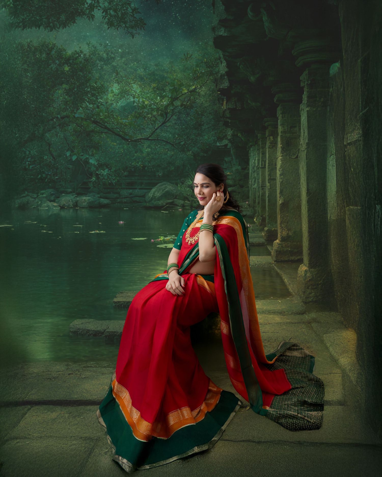 Woman in red and green saree sitting gracefully by ancient temple columns near a serene green lake.