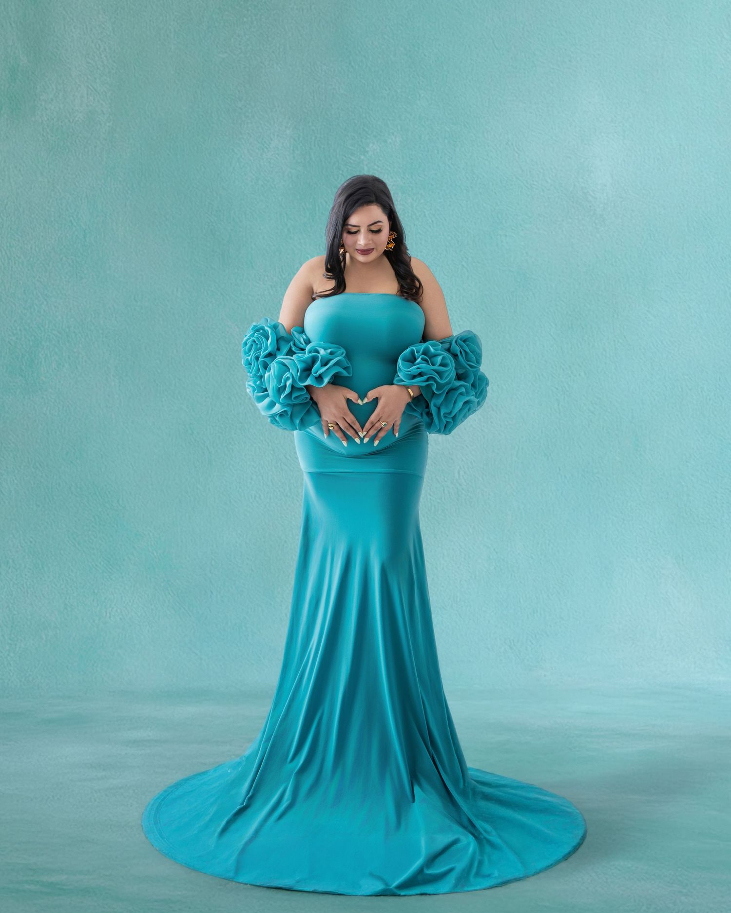Pregnant woman in elegant teal off-shoulder gown with ruffled sleeves, posing gracefully against a matching turquoise backdrop.