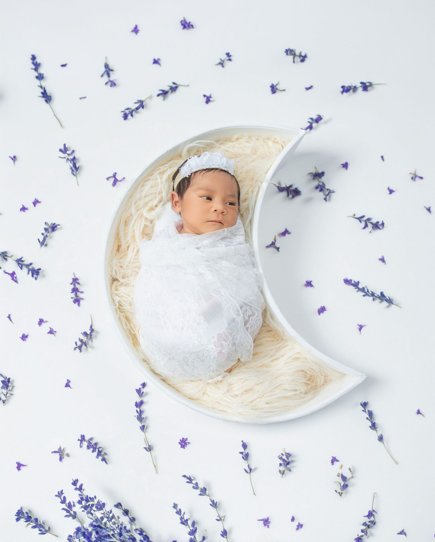 Newborn baby wrapped in white lace swaddle lying in a moon-shaped bowl surrounded by purple lavender flowers.