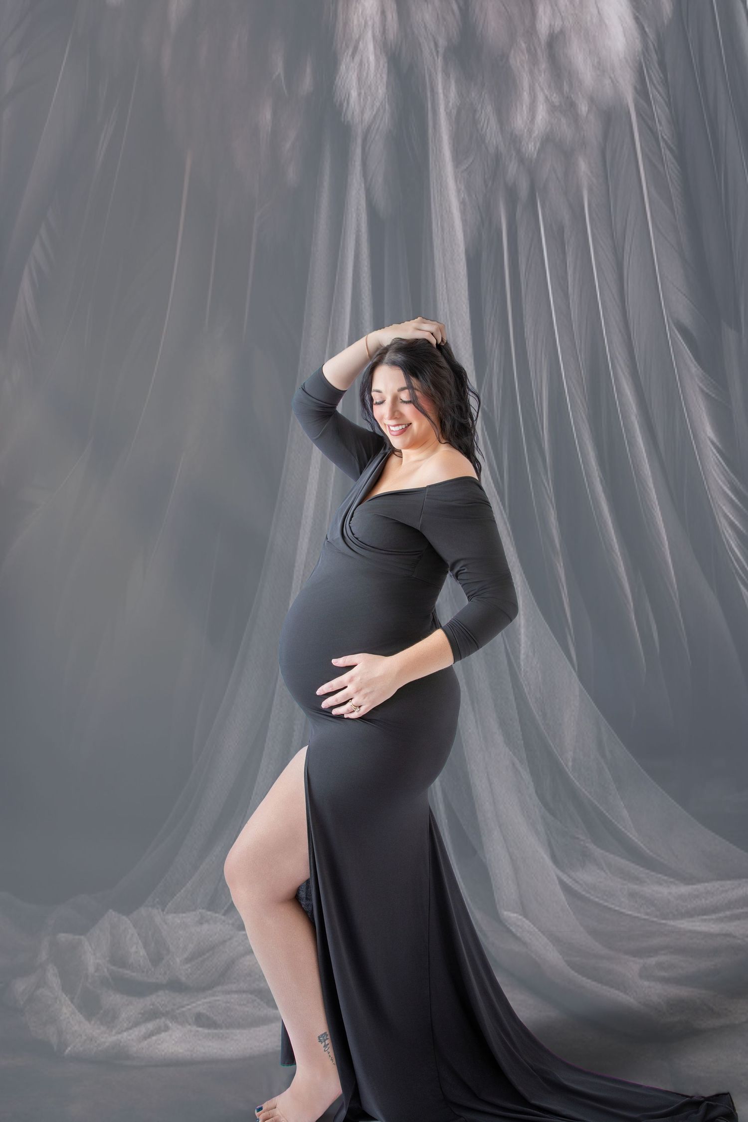 Pregnant woman in elegant black off-shoulder gown poses gracefully in studio with flowing gray fabric backdrop.