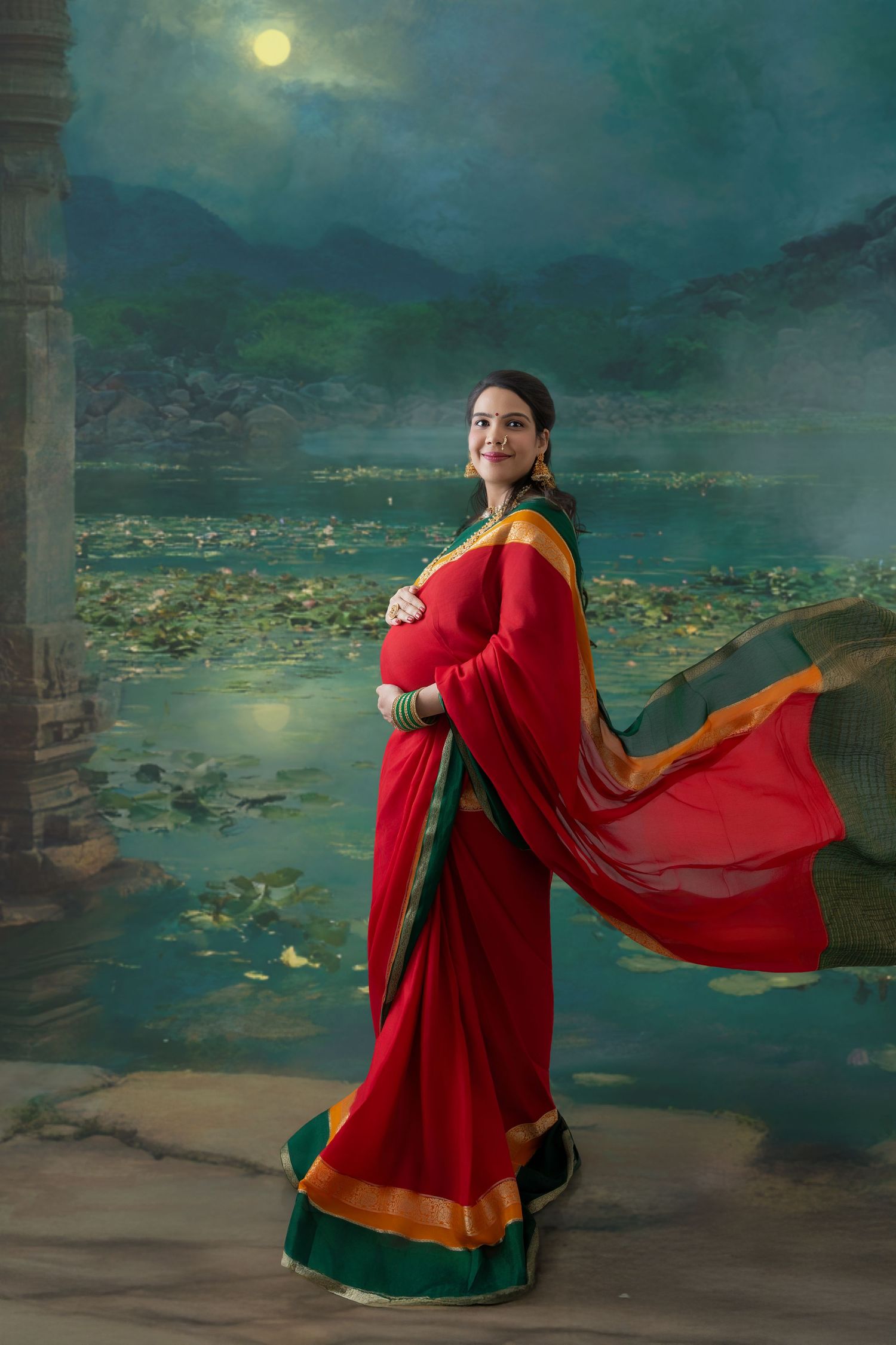 Pregnant woman in red saree with gold border poses gracefully against moonlit lake backdrop with lotus flowers.
