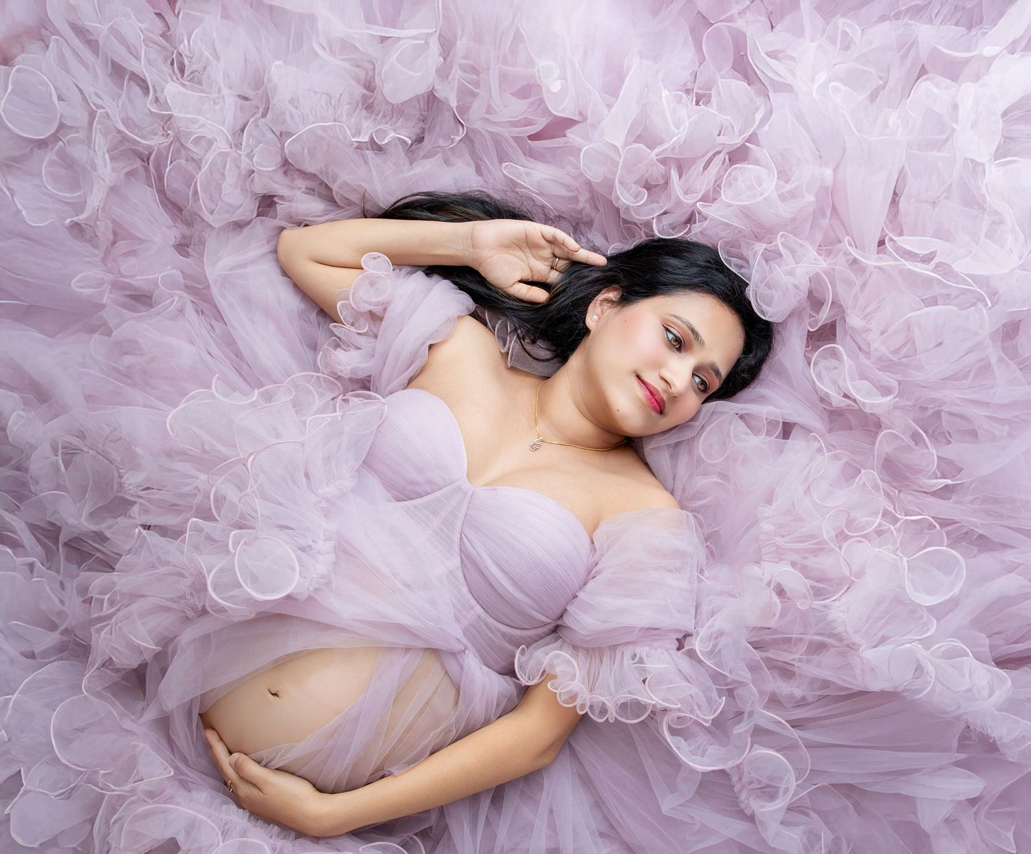 Pregnant woman in lavender tulle gown lying down, maternity photography session with elegant pink ruffled dress.