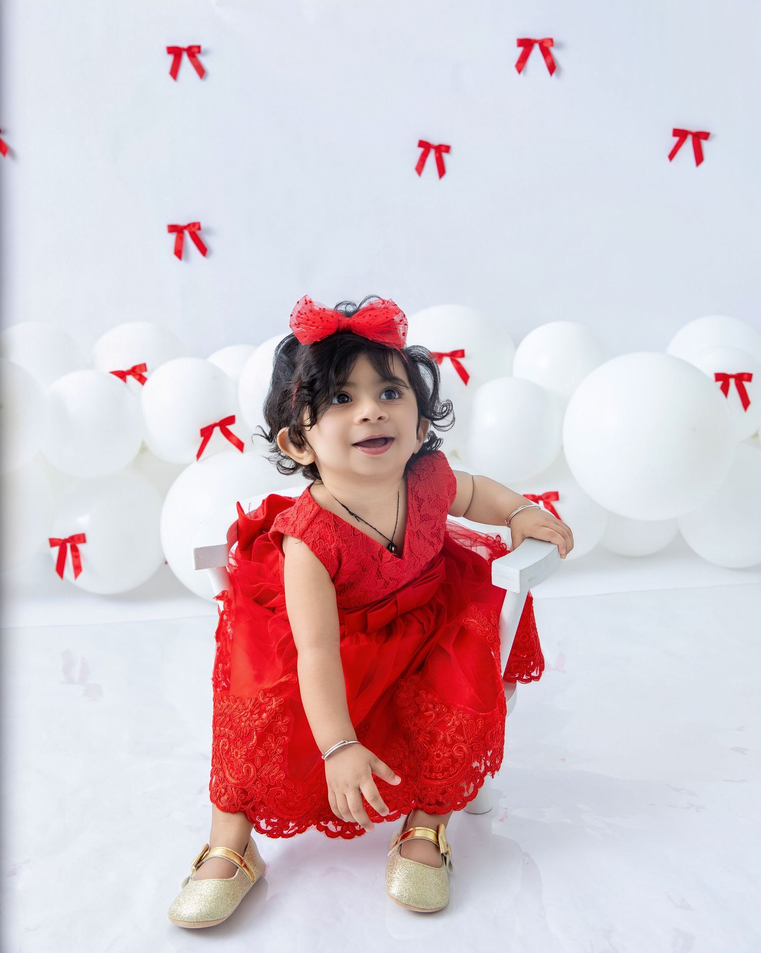 A toddler in a red dress and gold shoes crouches on a white floor with decorative bows.