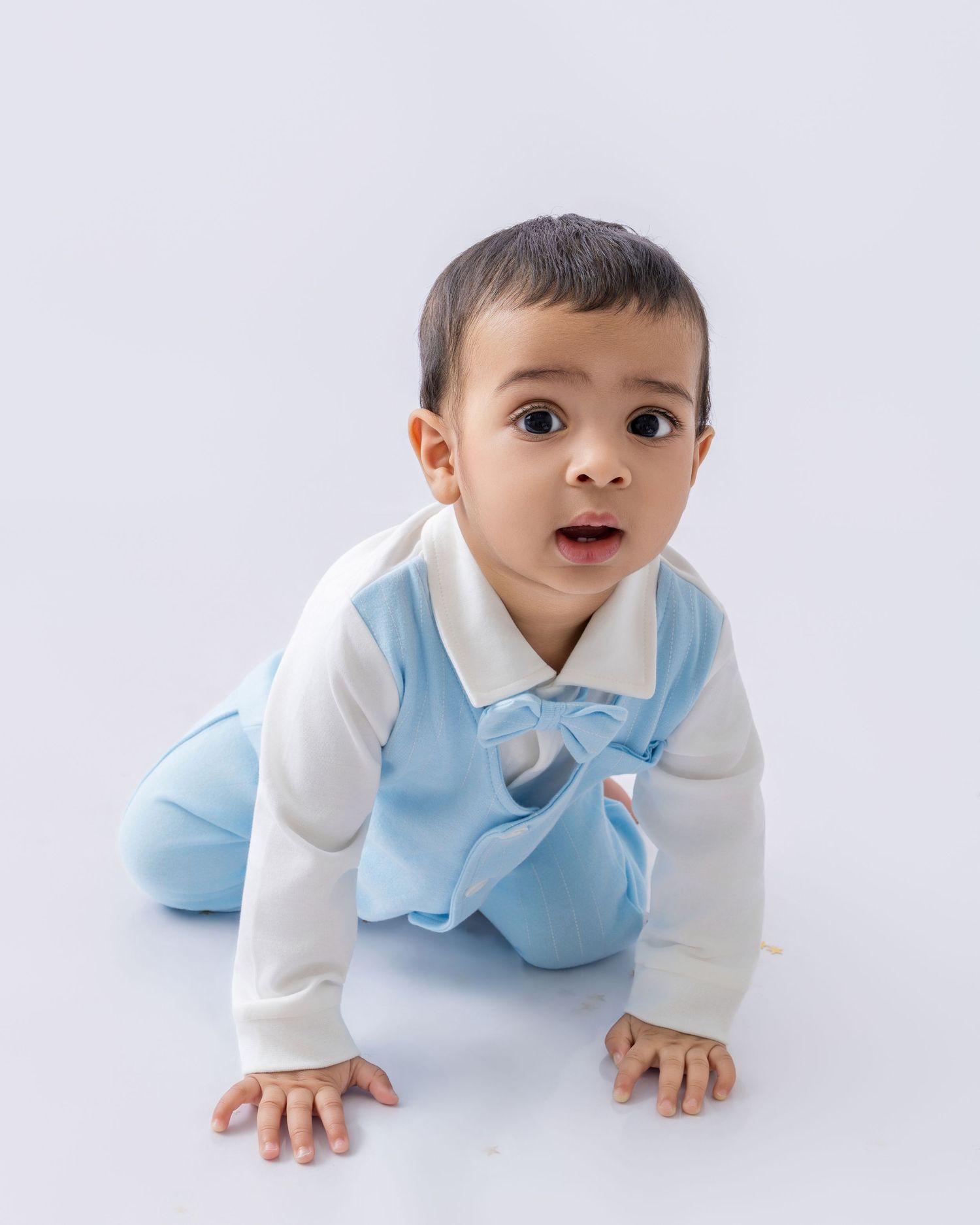 Baby in light blue and white outfit crawls on white backdrop during professional photo session.