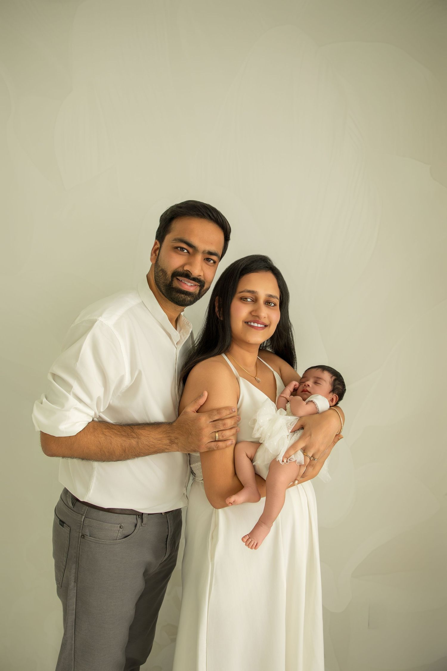 Parents in coordinated white and gray attire cradle their infant during an intimate photoshoot against a neutral wall.