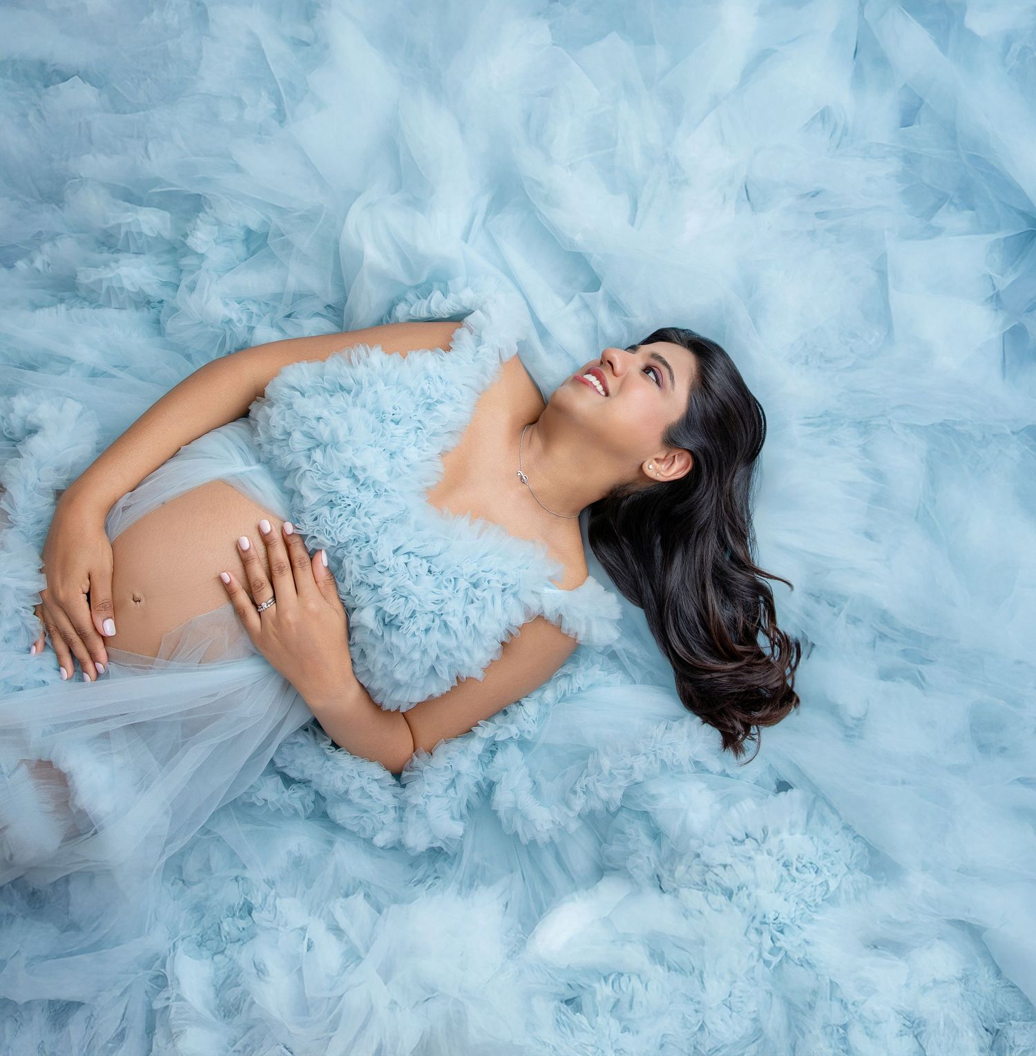 Woman in flowing light blue tulle gown lying gracefully on cloud-like fabric background.