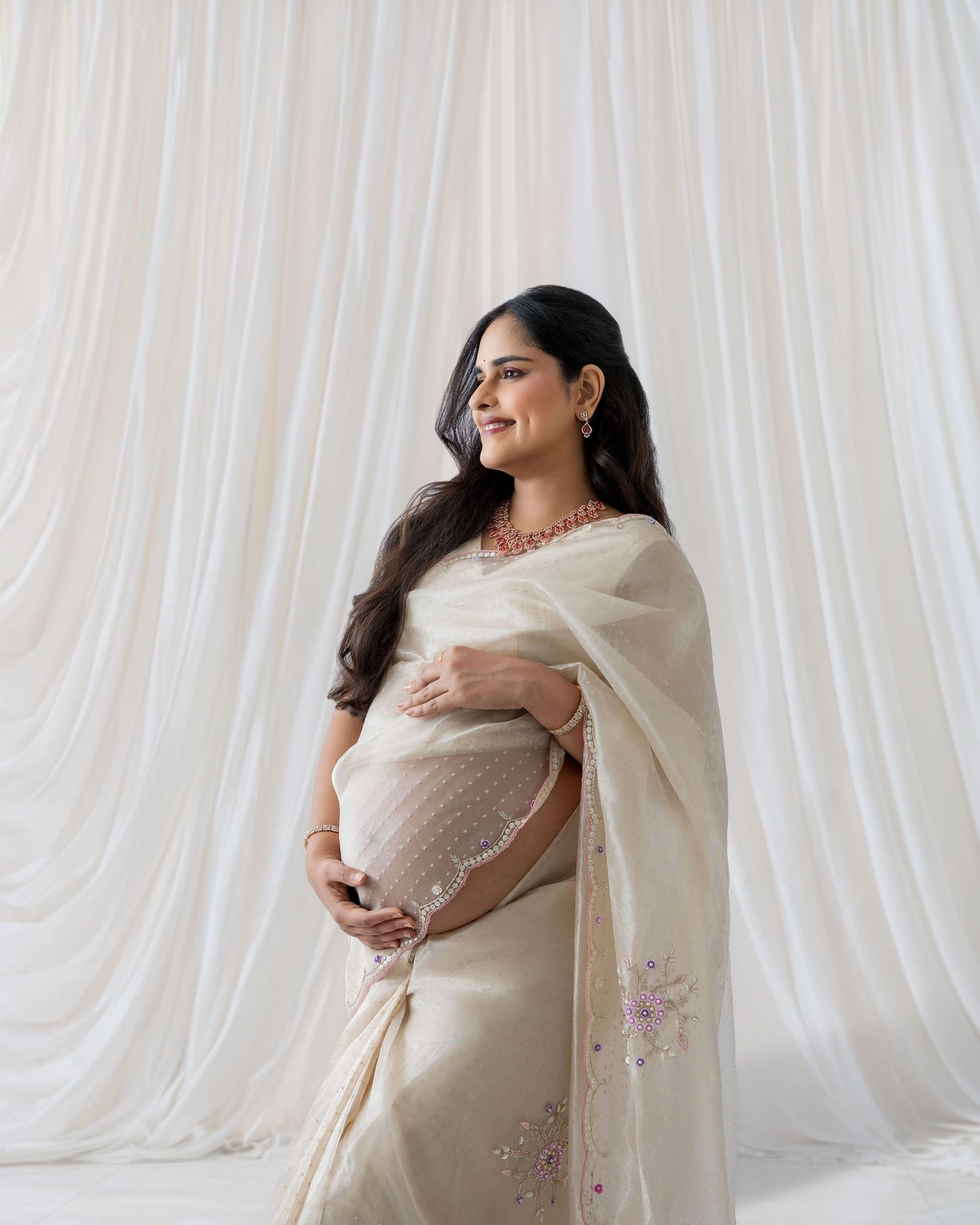 Smiling pregnant woman in cream floral saree with ruby necklace stands elegantly against white curtain background.