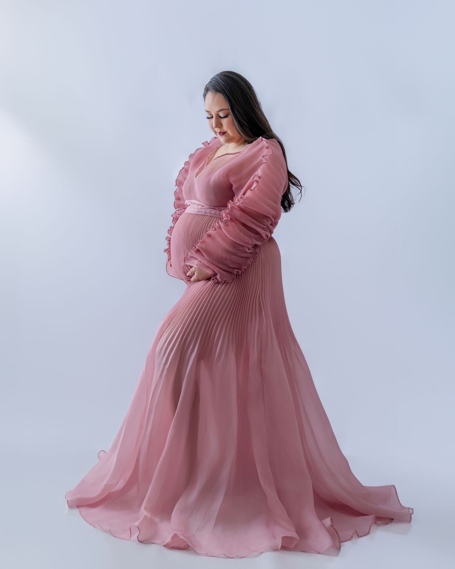 Pregnant woman in elegant dusty pink pleated maternity gown with long sleeves, posing gracefully against white background.
