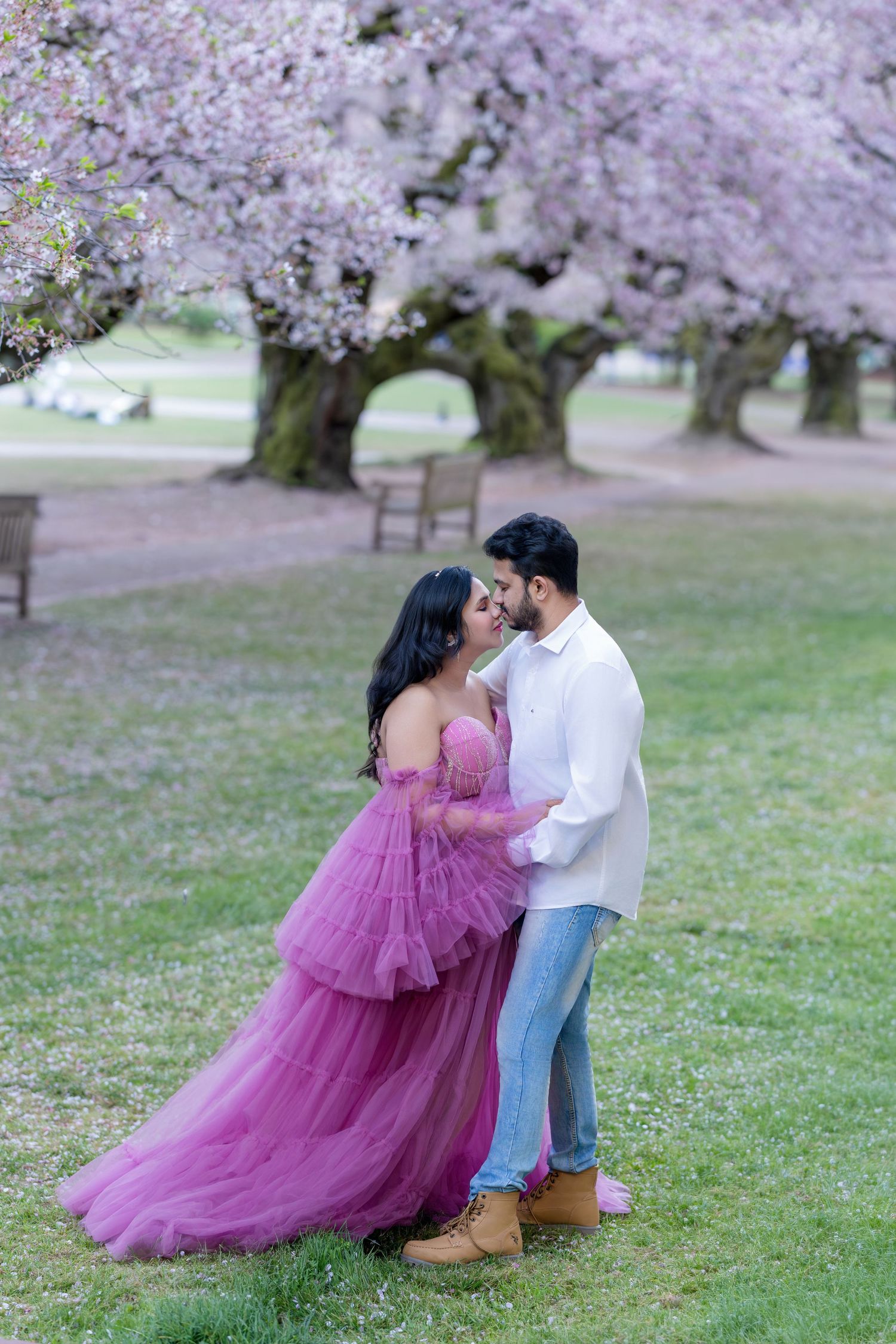 Couple embracing under blooming cherry blossom trees, woman in purple tulle gown, man in white shirt and jeans.