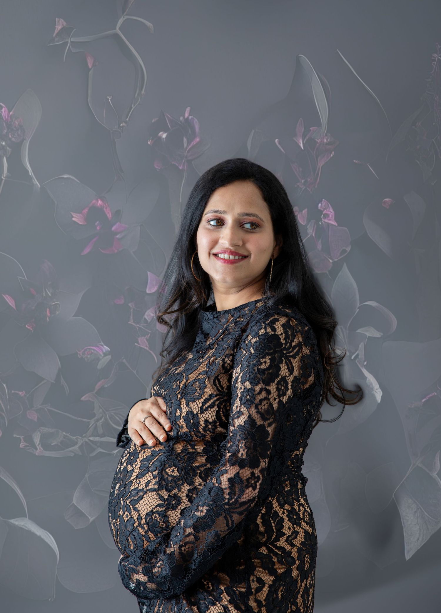 Pregnant woman in black lace dress posing sideways against gray floral background.