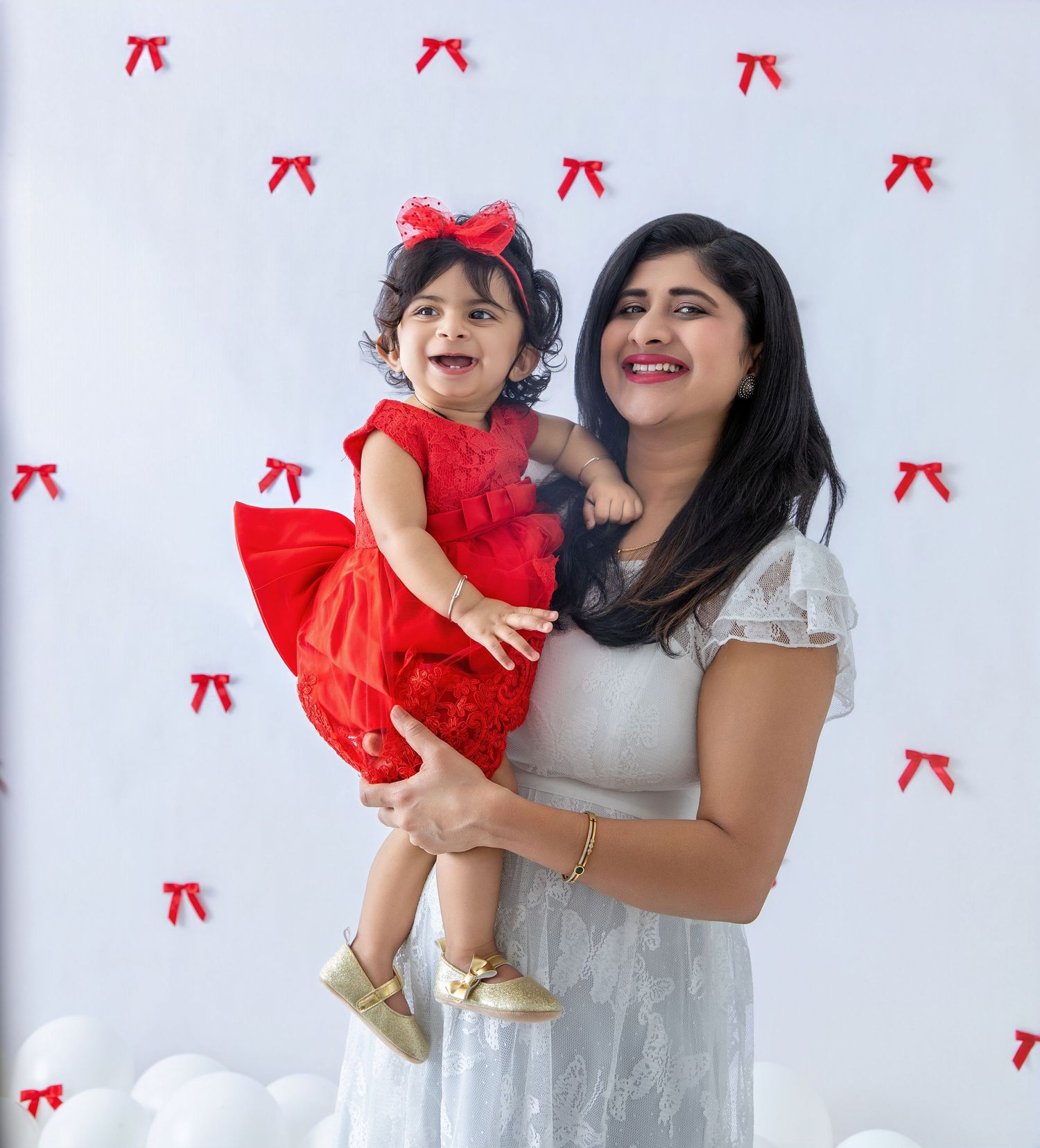 Mother and child in white and red dresses smile and laugh against wall with red bow decorations.