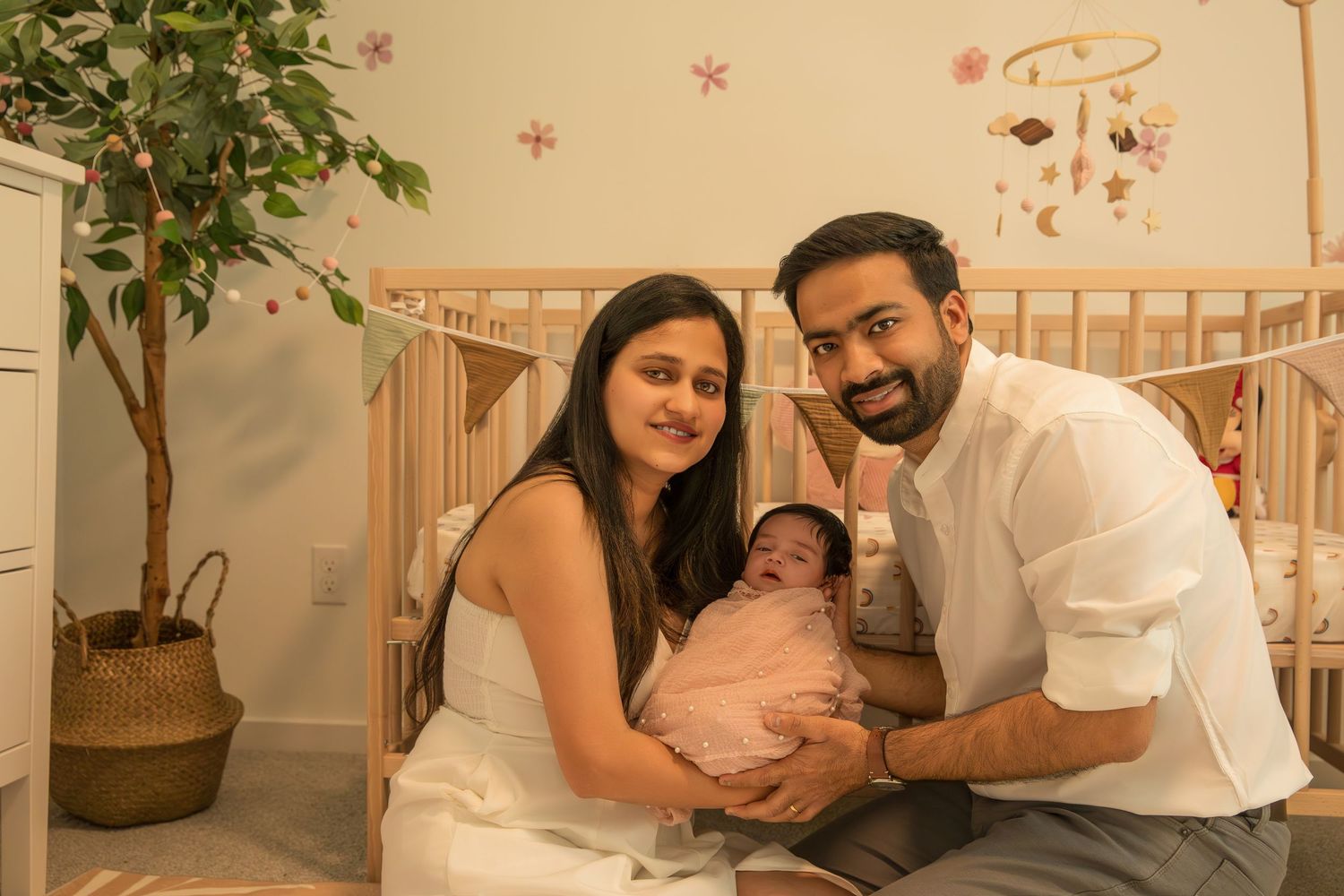 Parents sitting in nursery room with potted plant and crib while holding their newborn baby.