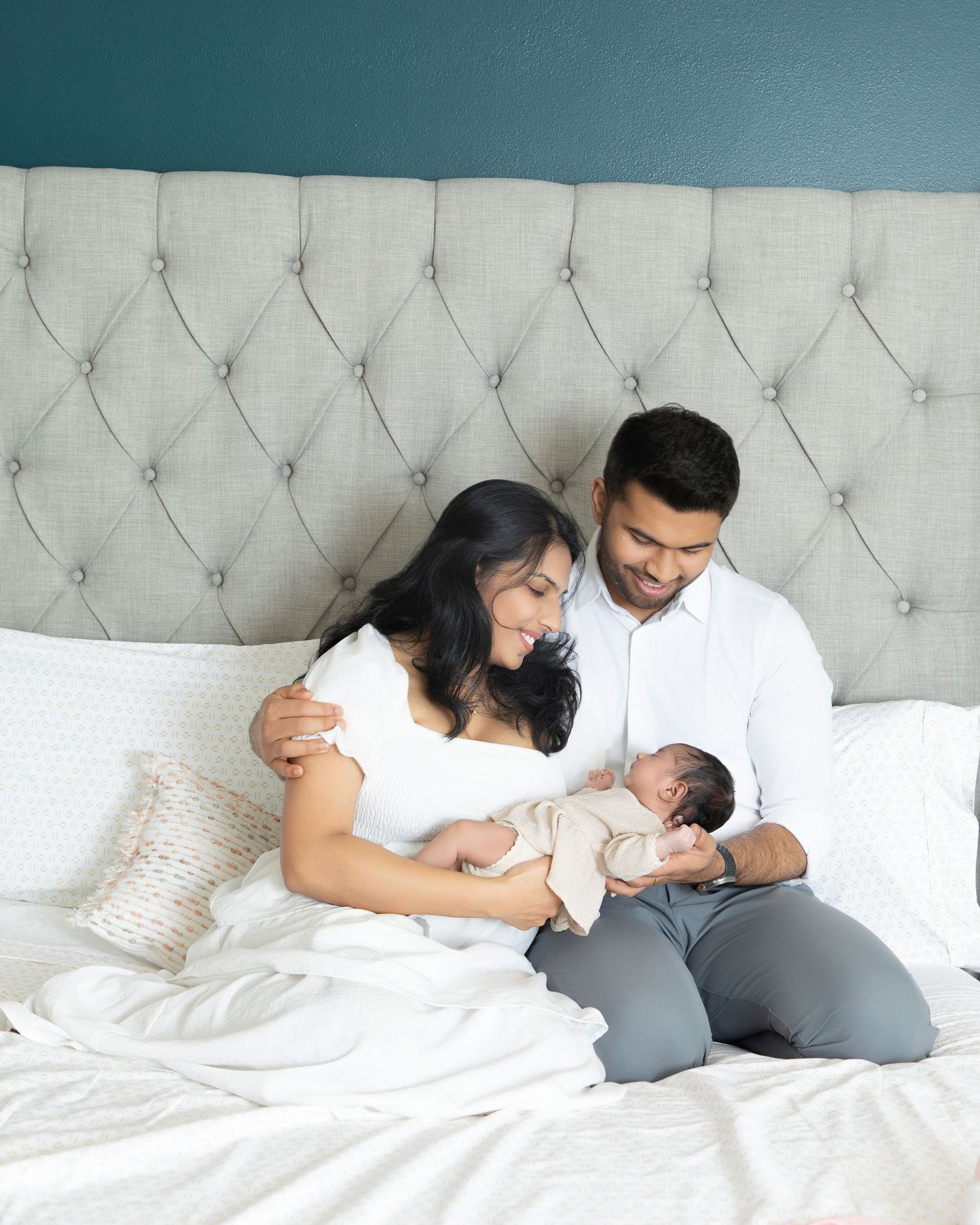 Parents sitting on bed with newborn baby wrapped in white blanket against tufted headboard.